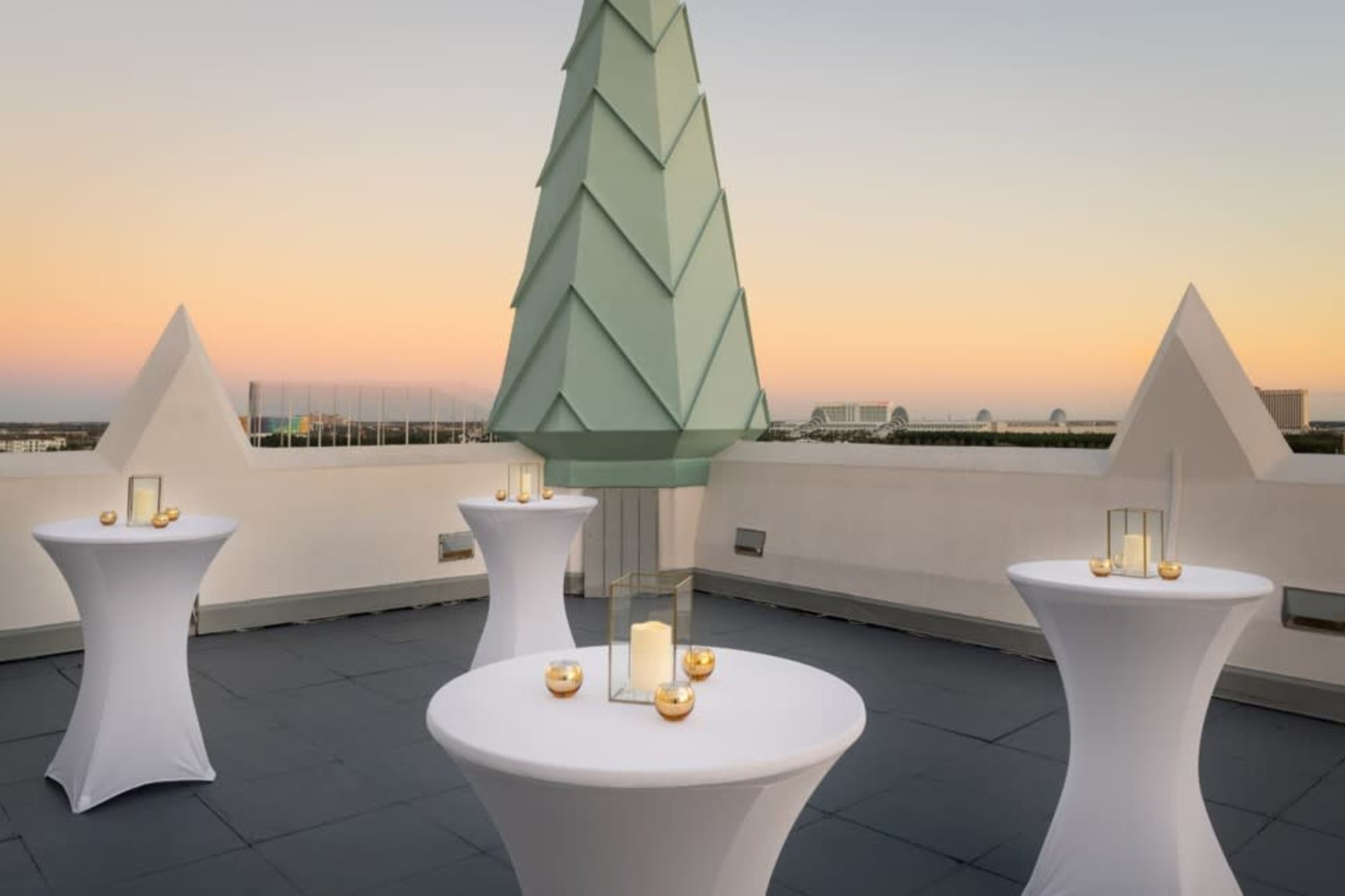 The image shows a rooftop event space with white tables surrounded by decorative candle holders against a sunset sky.