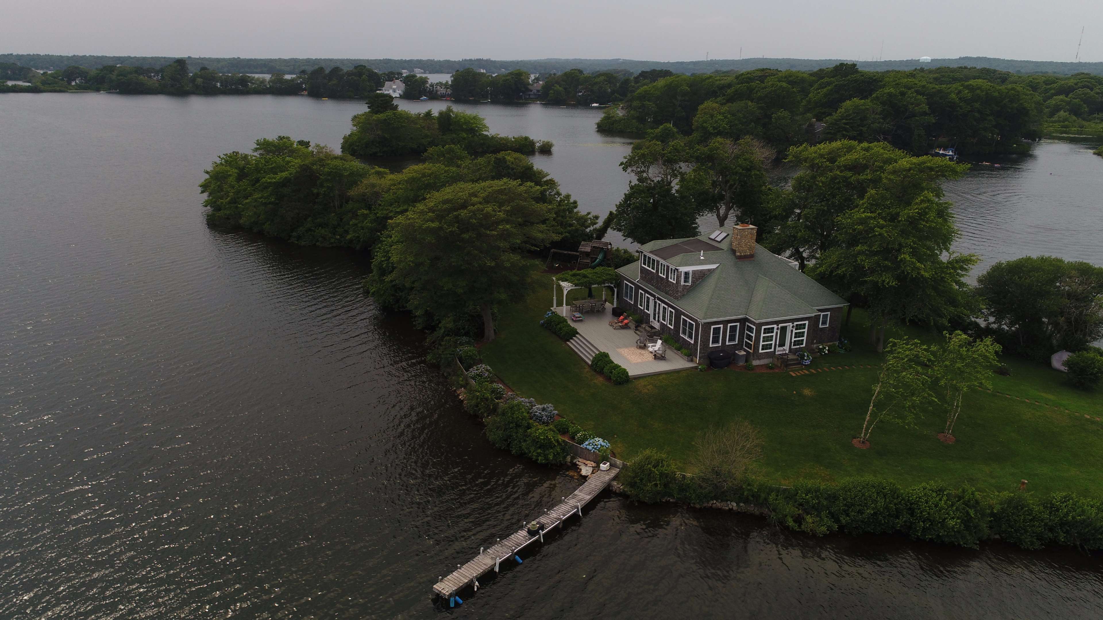 A large house with a green roof is situated on a small island surrounded by water, featuring a dock extending into the lake.