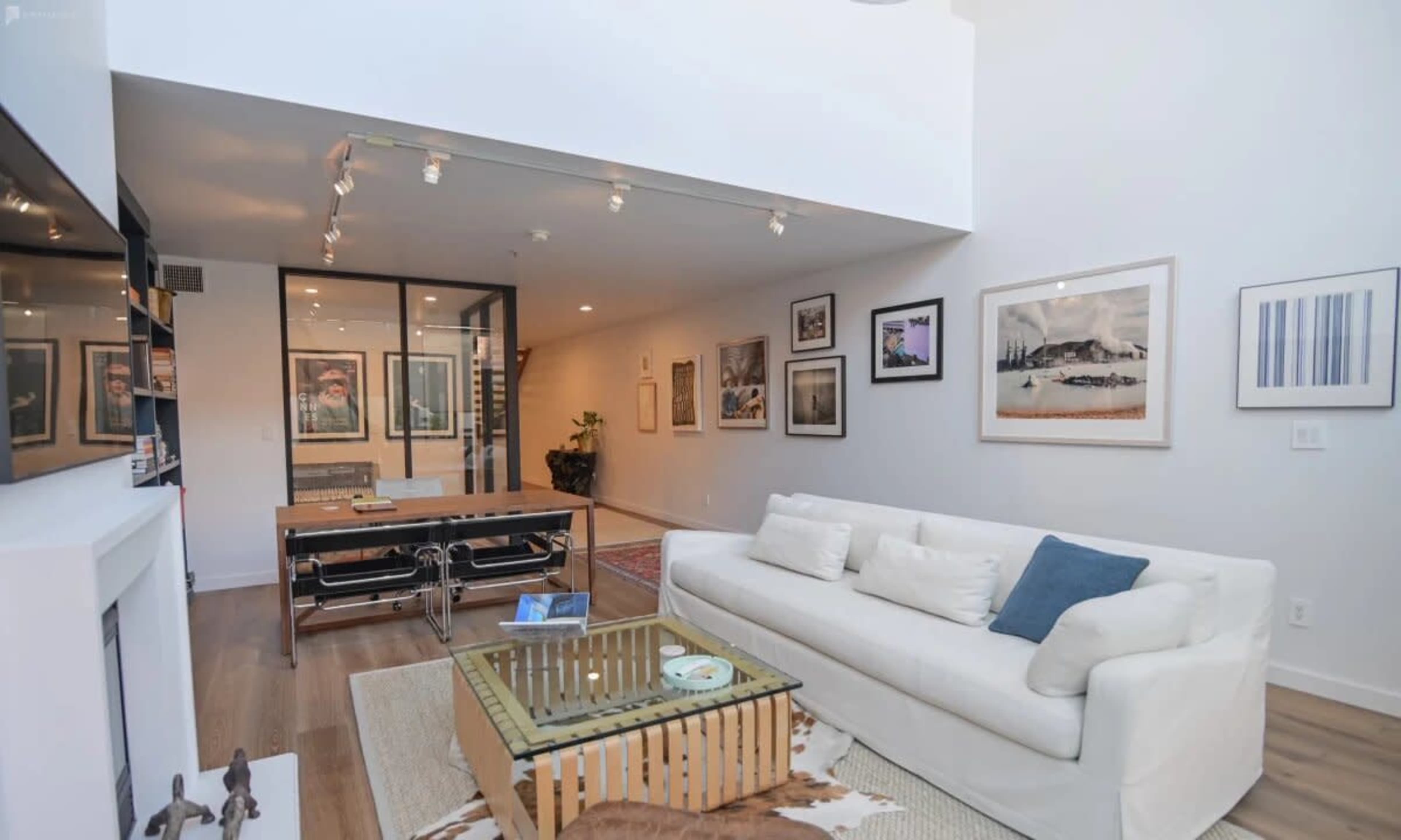 A modern living room with a white sofa, a glass coffee table, and several framed artworks on the walls.
