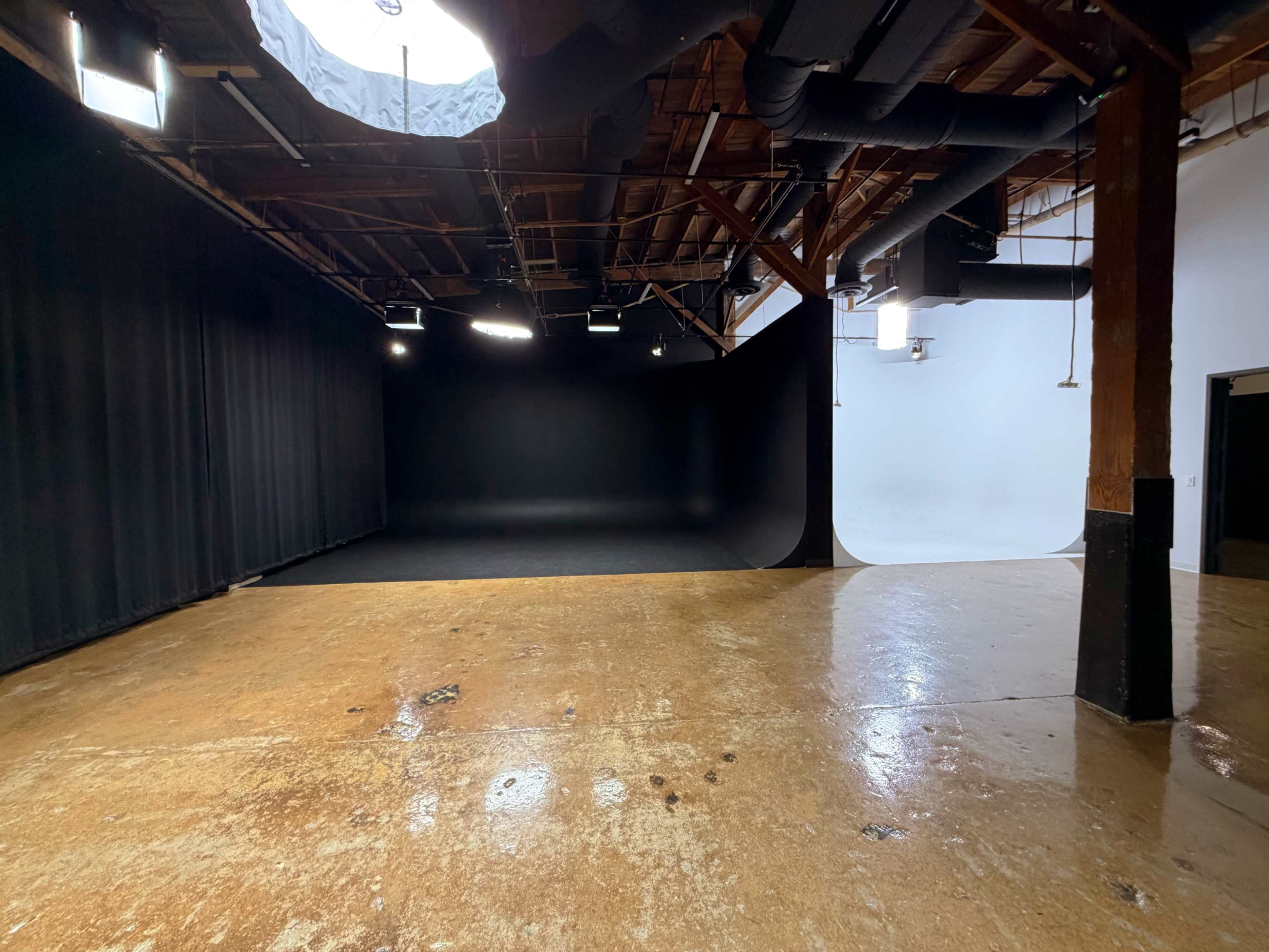 The image shows an empty studio space featuring a black backdrop on one side and a white backdrop on the other, with polished concrete flooring and overhead lighting.