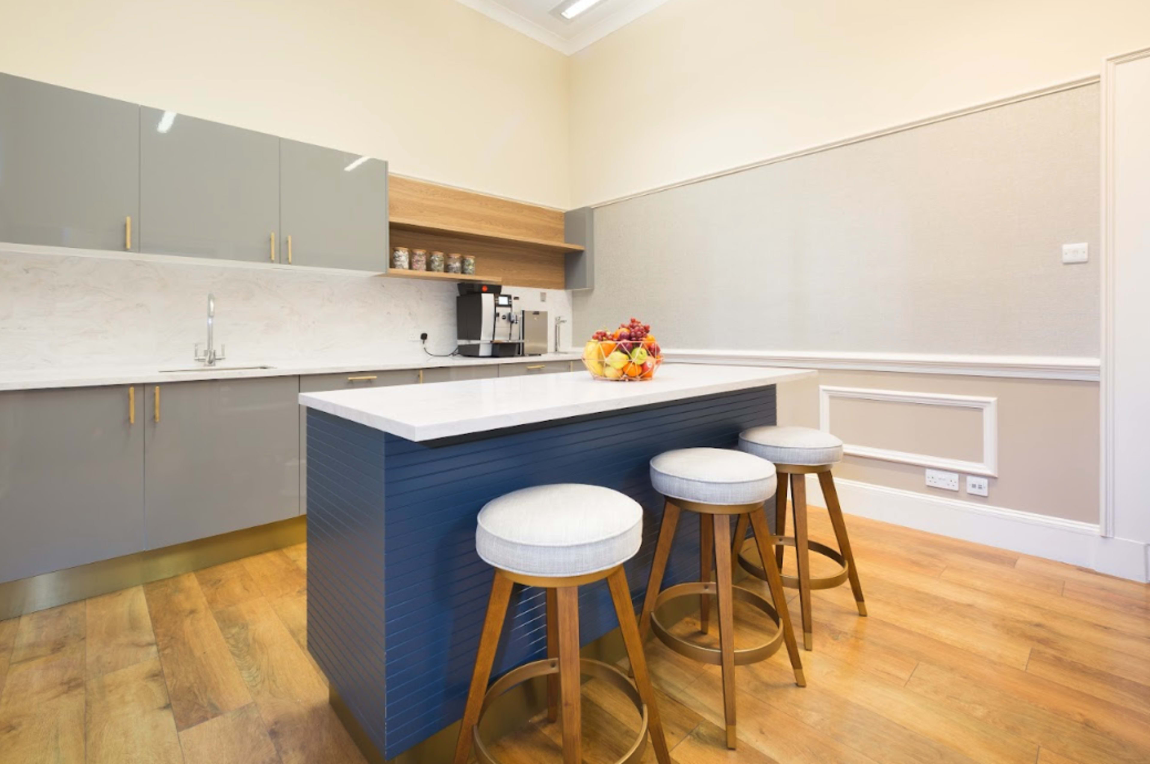 A modern kitchen features gray cabinets, a marble backsplash, a wooden countertop, and three bar stools at an island with a bowl of fruit.