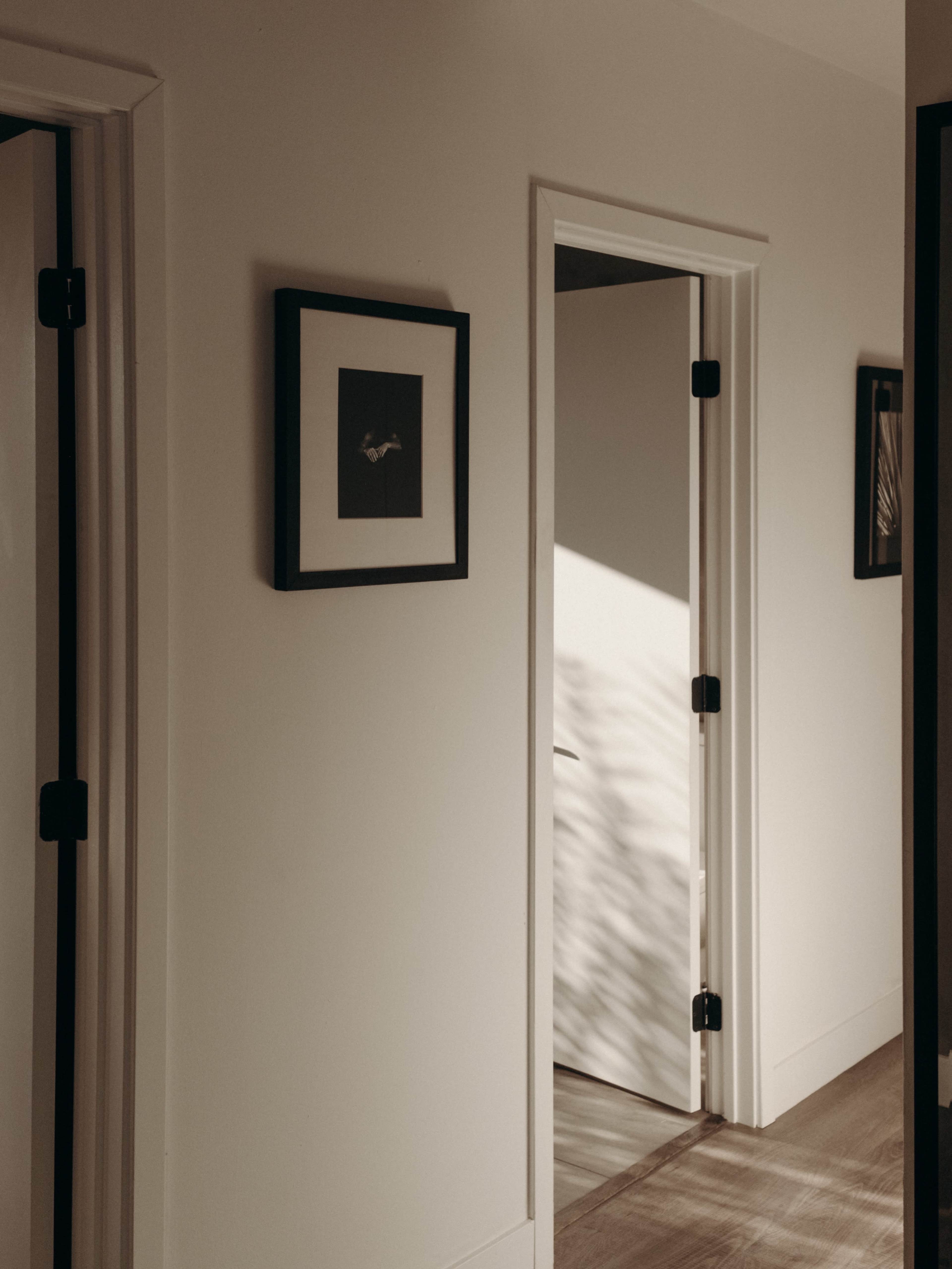 A narrow hallway features two doors, a framed picture on the wall, and soft shadows cast on the floor.
