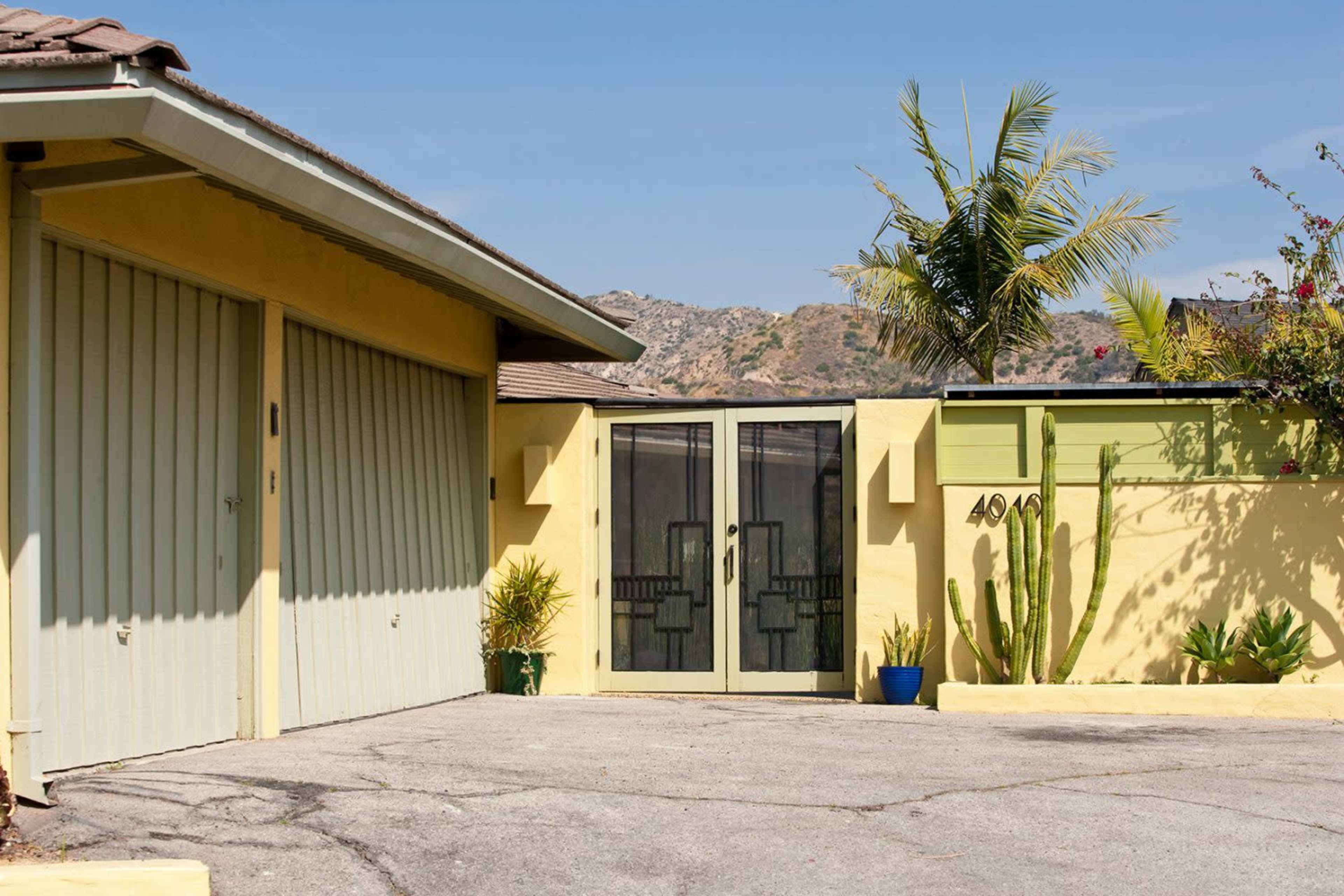 The image shows a yellow building with a double door entrance, flanked by plants and a driveway.