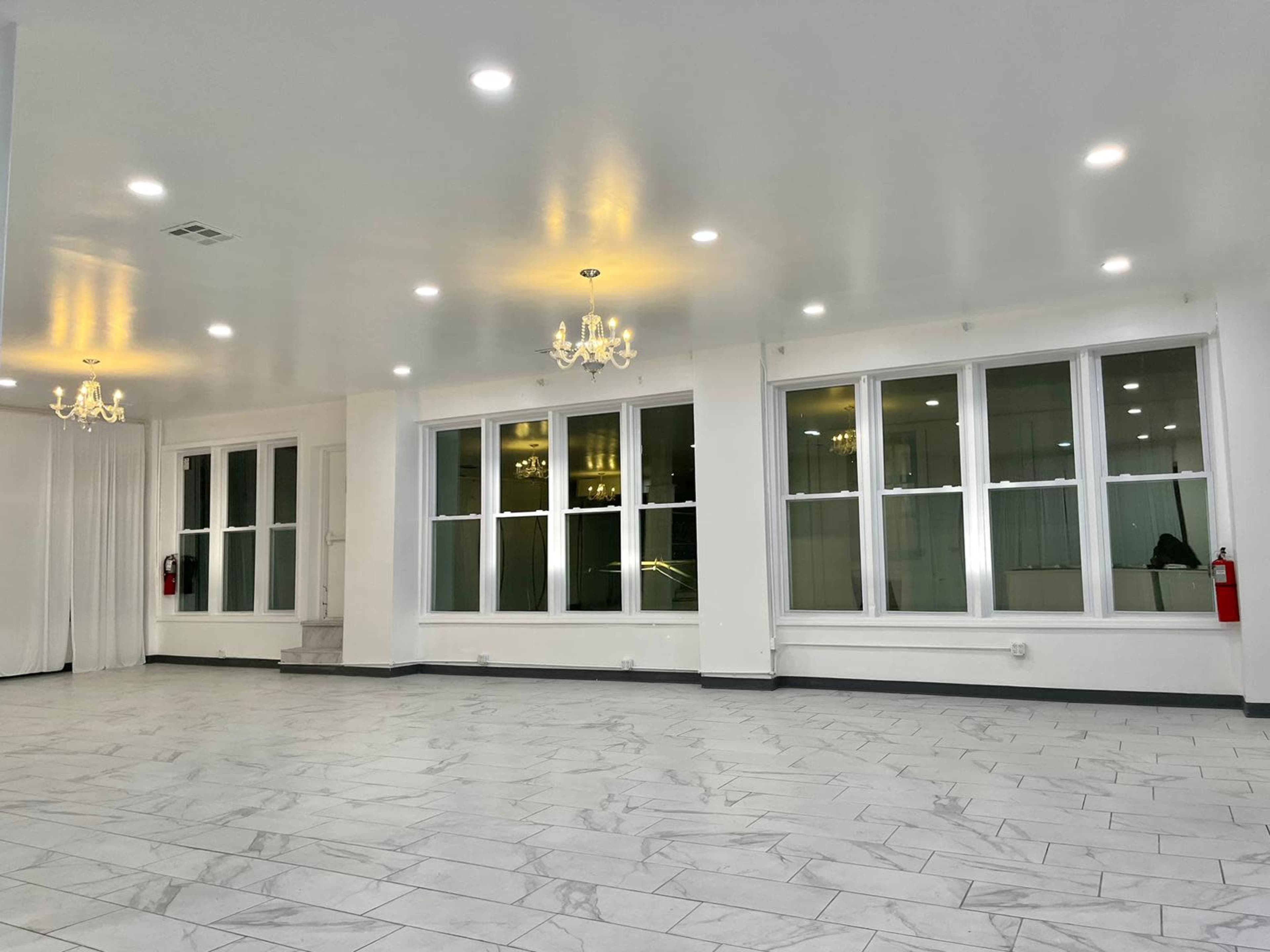 An empty room with white walls, multiple large windows, and chandeliers, featuring a tiled floor.