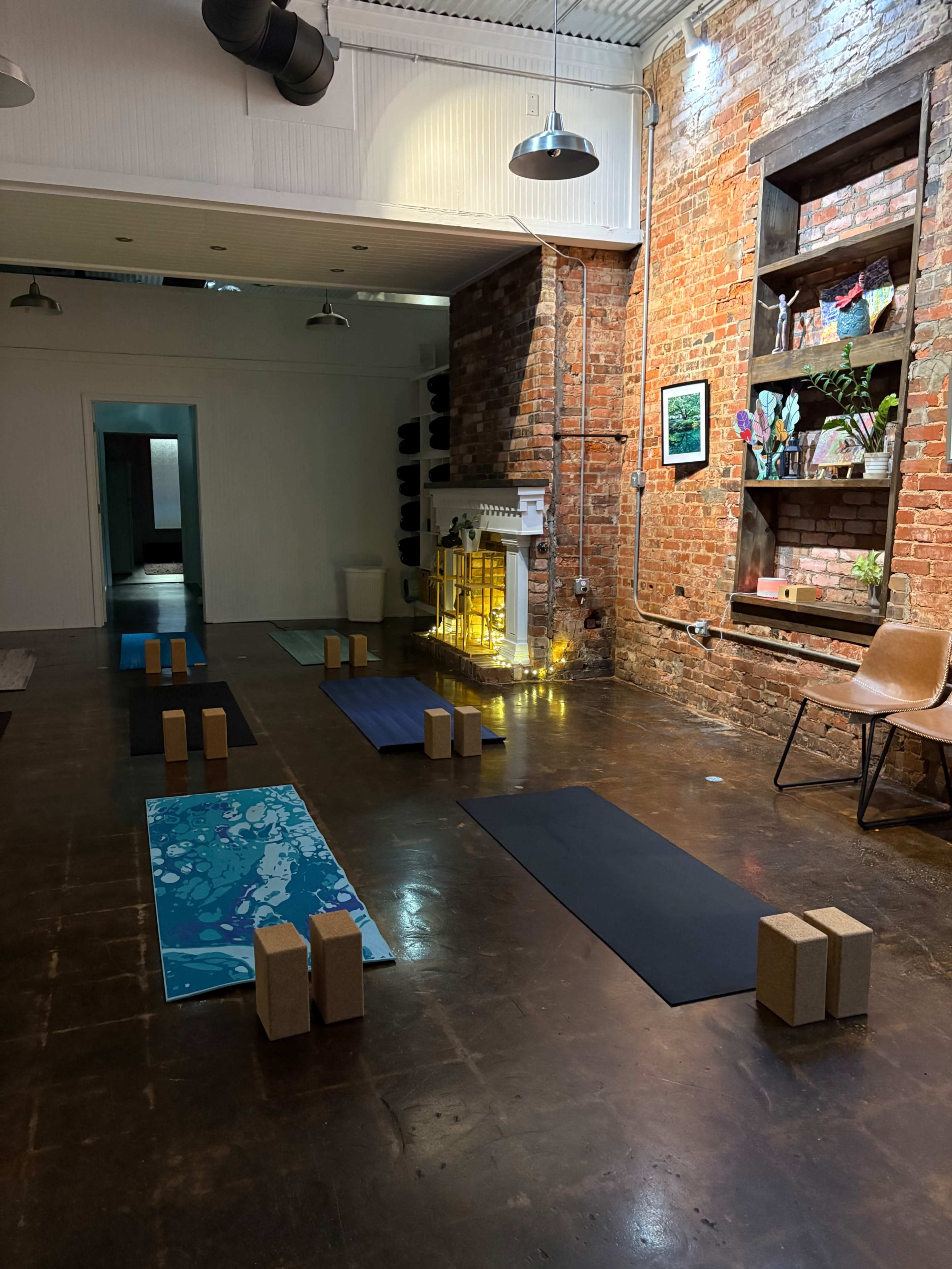 The image shows a yoga studio featuring several yoga mats arranged on a dark floor, with cork blocks placed at intervals and brick walls adorned with shelves and decorative items.