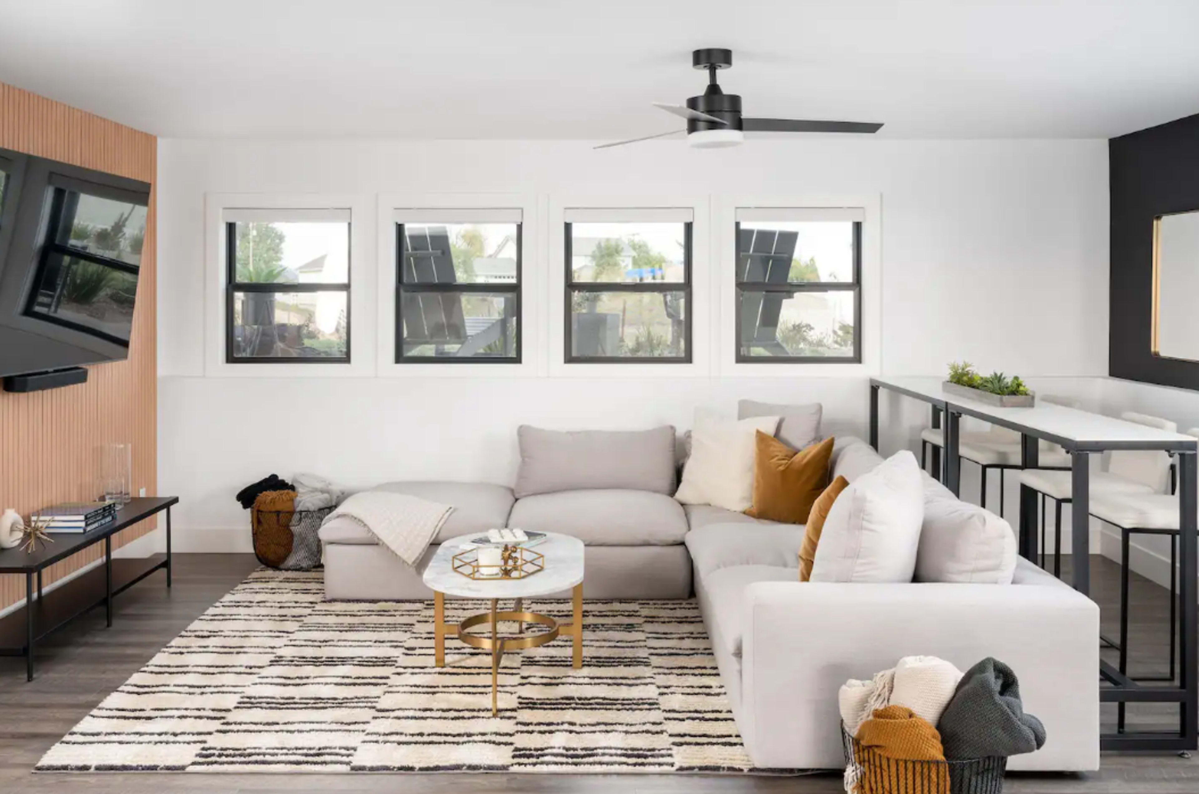 A modern living room featuring a light gray sectional sofa, a round coffee table, and a striped area rug, with large windows allowing natural light to enter.