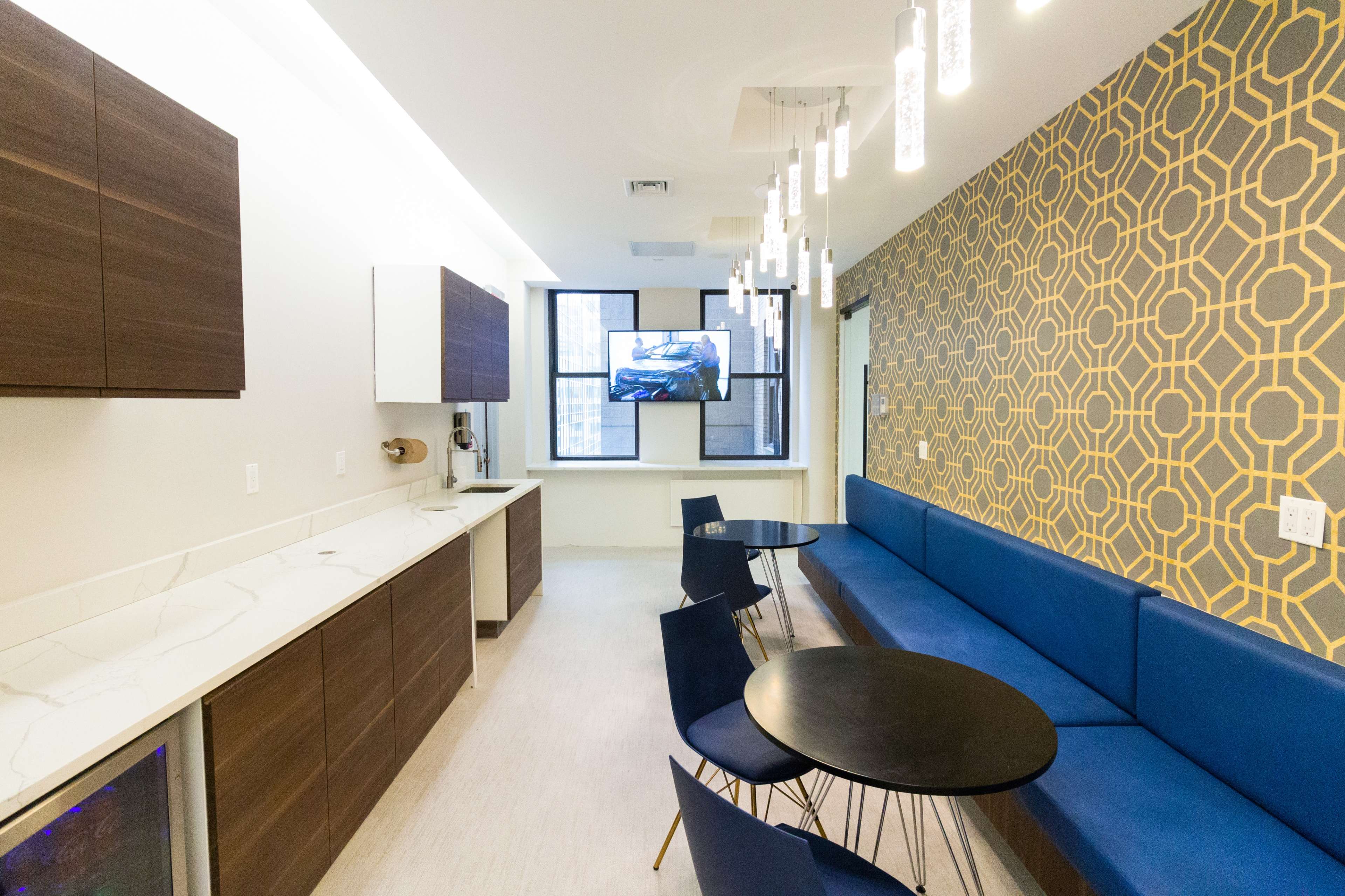 A modern kitchenette area with a long blue couch, round black tables, and a television mounted on the wall.