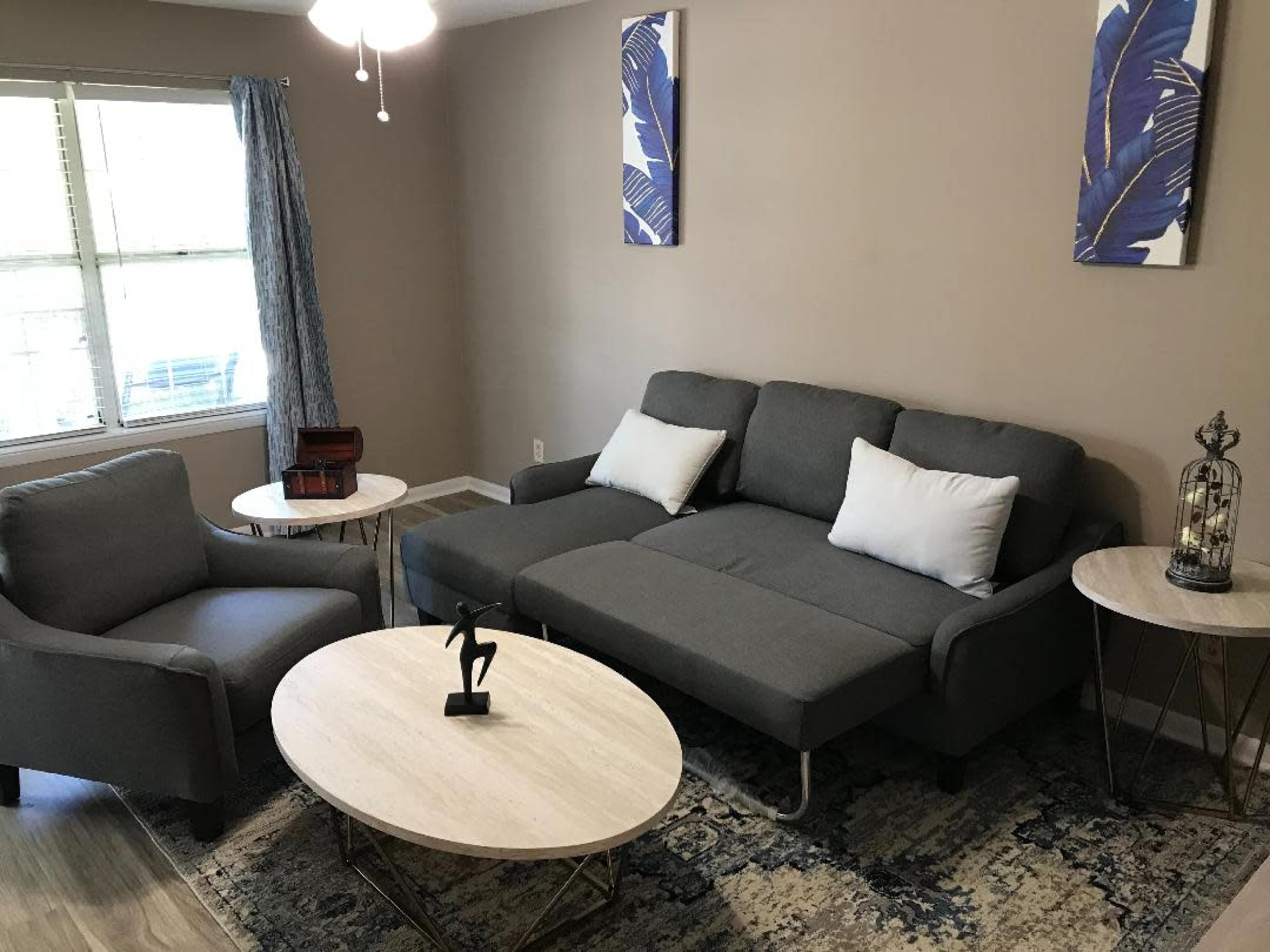 A neatly arranged living room with a gray sectional sofa, two accent chairs, and two coffee tables on a patterned rug, under a ceiling fan.