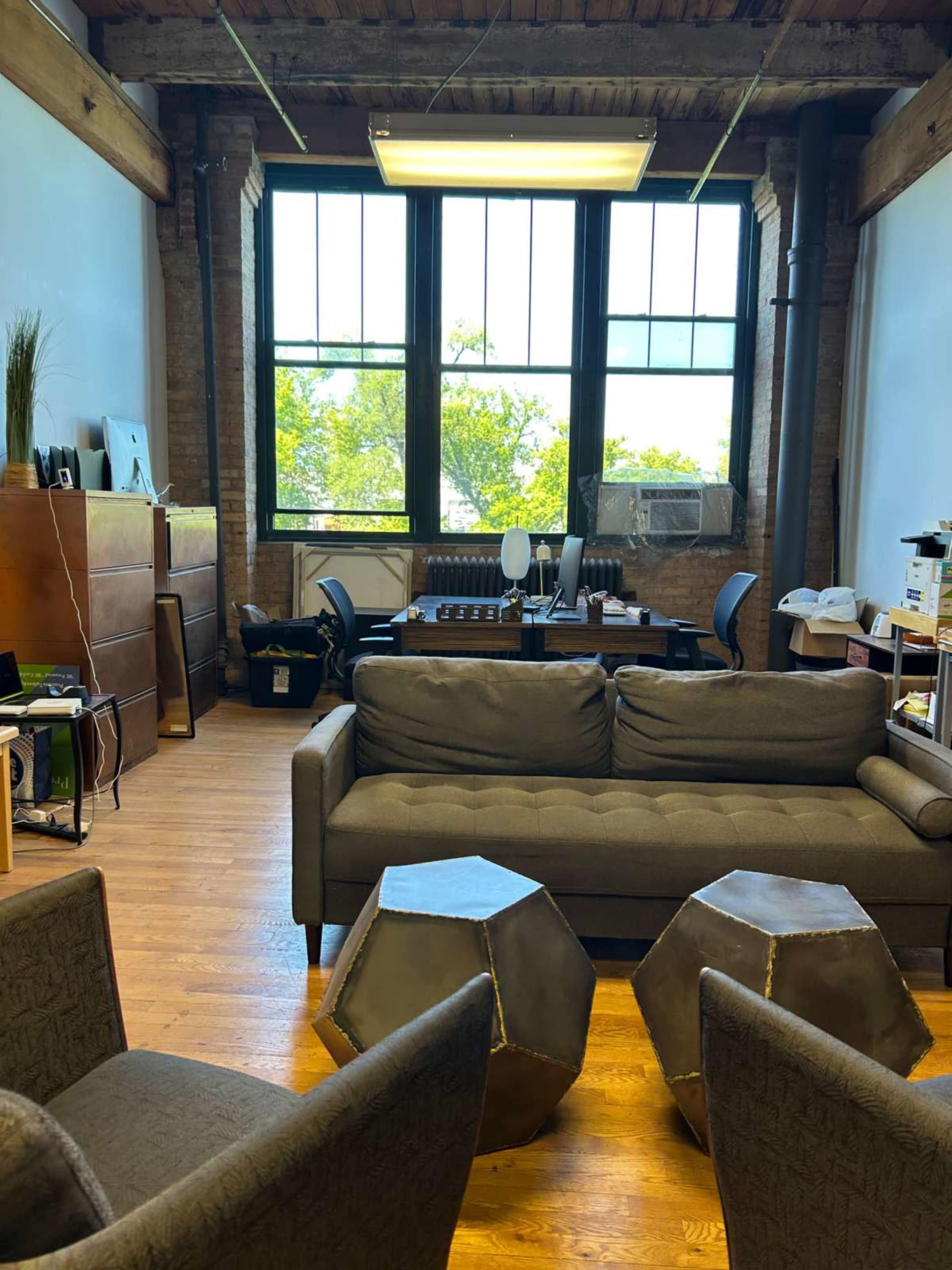 The image shows a workspace with a sofa, two armchairs, and a large window overlooking greenery.