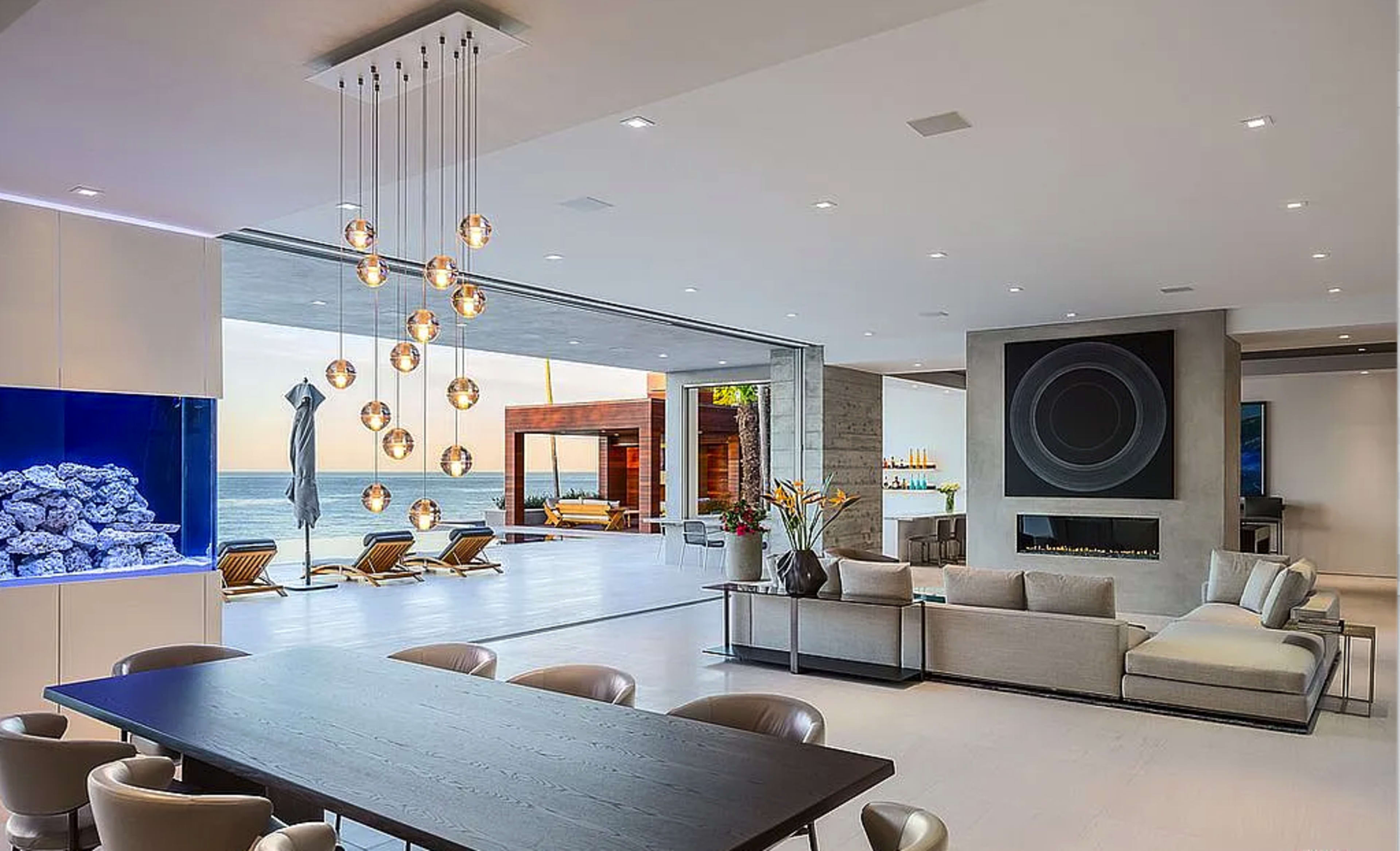 The image shows a modern open-concept living space with a dining area, a lounge section, and large glass doors overlooking an ocean view.