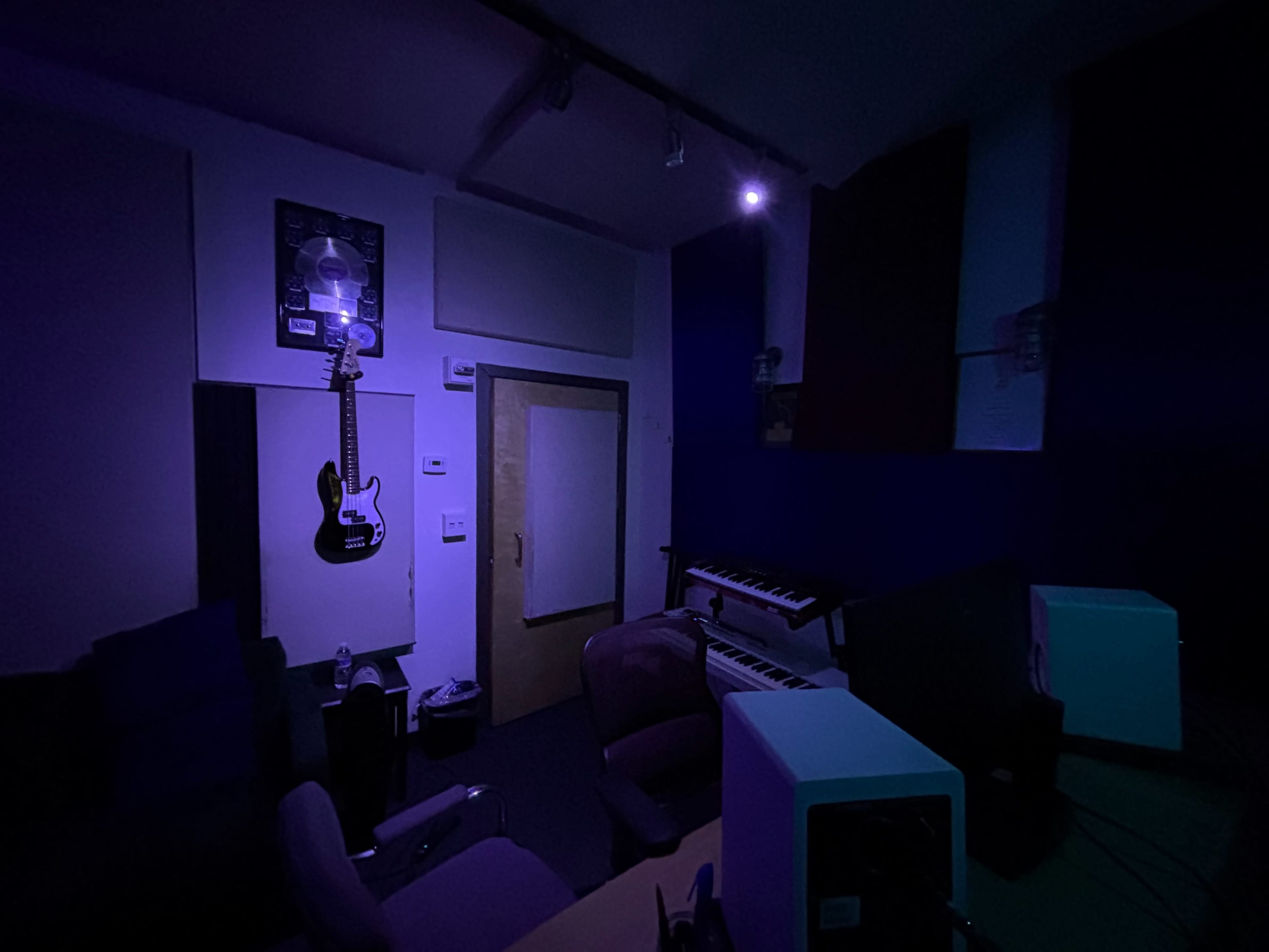 The image shows a dimly lit music studio featuring a black guitar hanging on the wall, a keyboard, and purple seating.