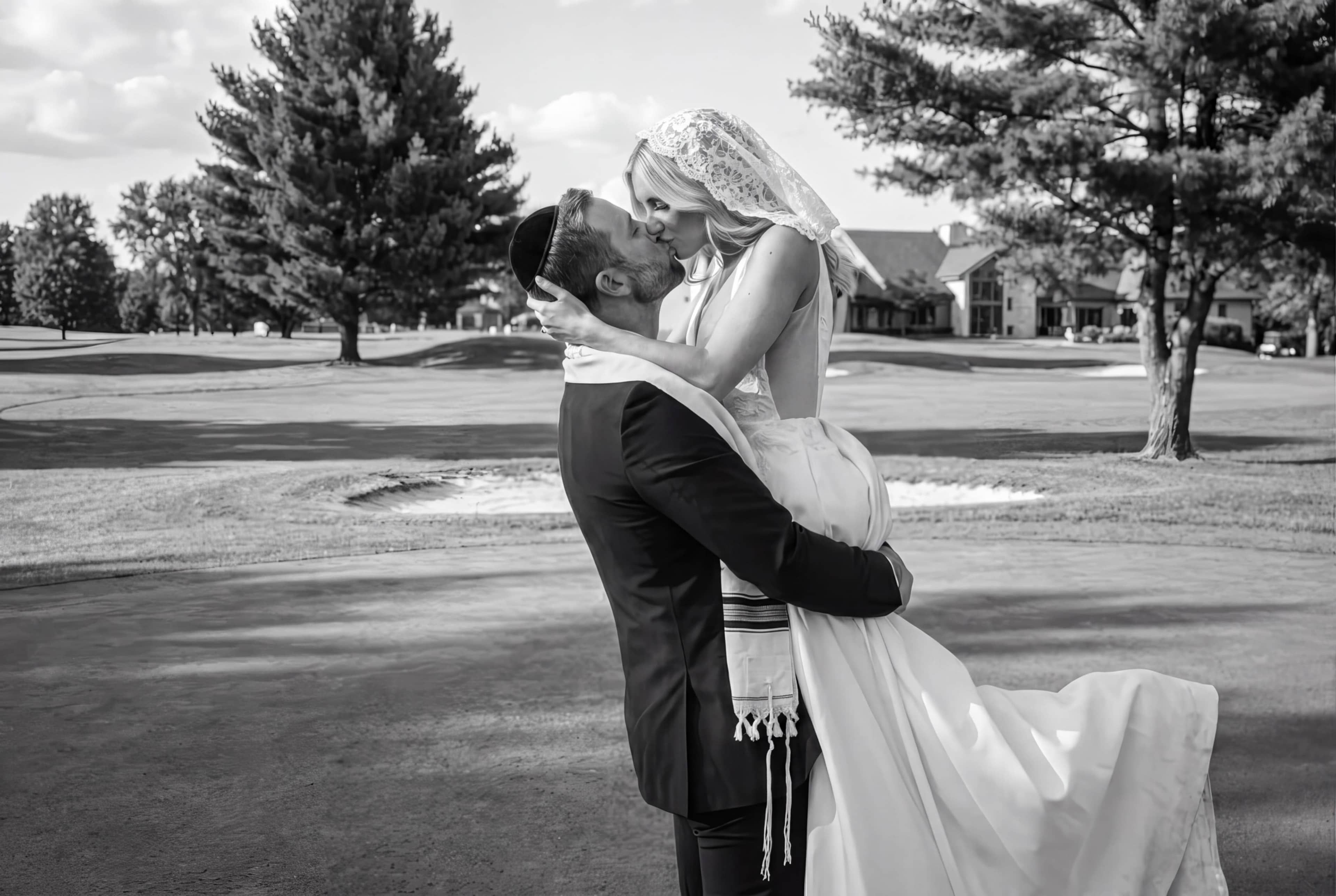 A groom in a black suit lifts his bride, who is wearing a white wedding dress and veil, in an embrace on a golf course.