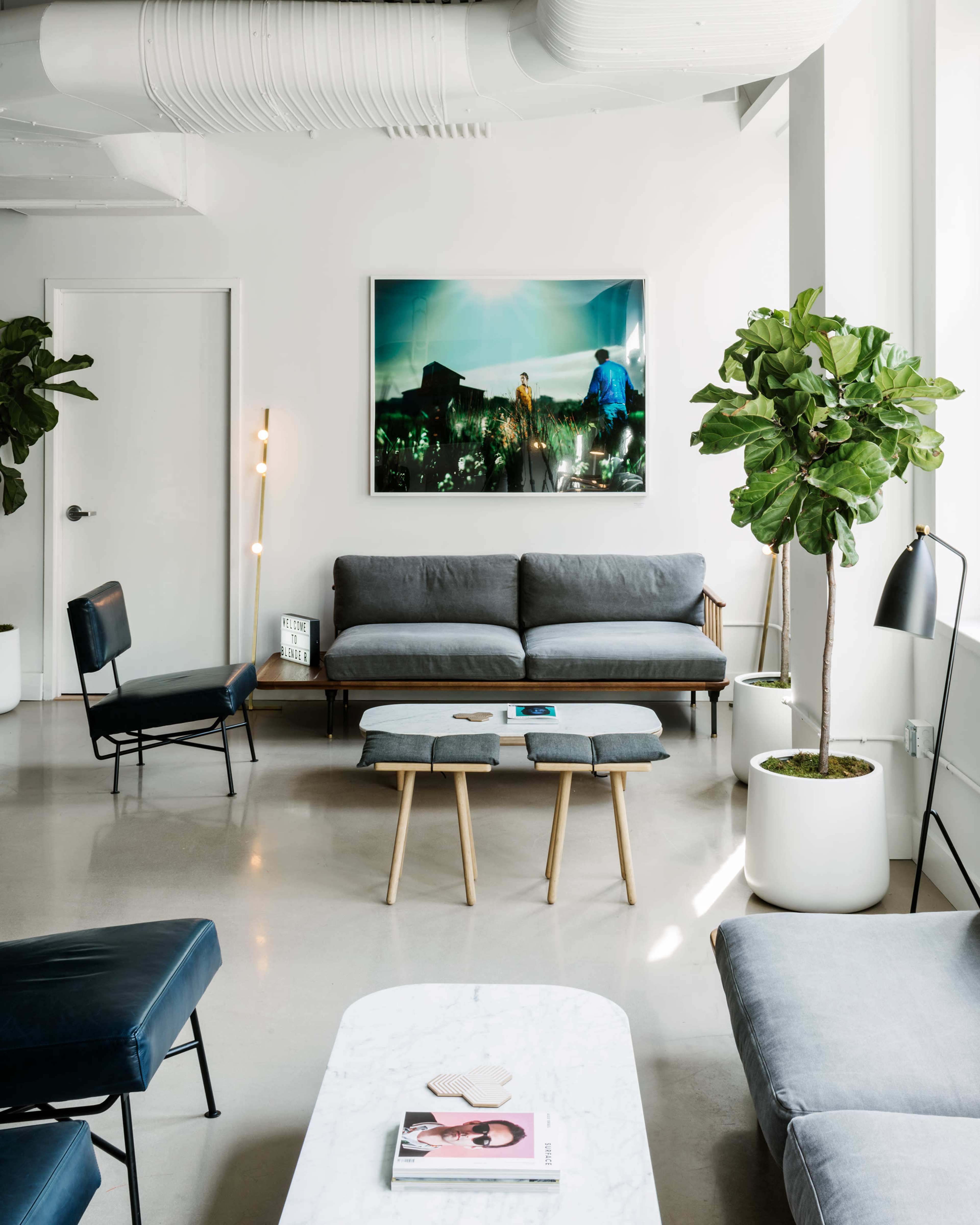 The image shows a modern, minimalistic living space featuring a gray sofa, a coffee table with two stools, and potted plants, all against a backdrop of large windows and a colorful artwork on the wall.