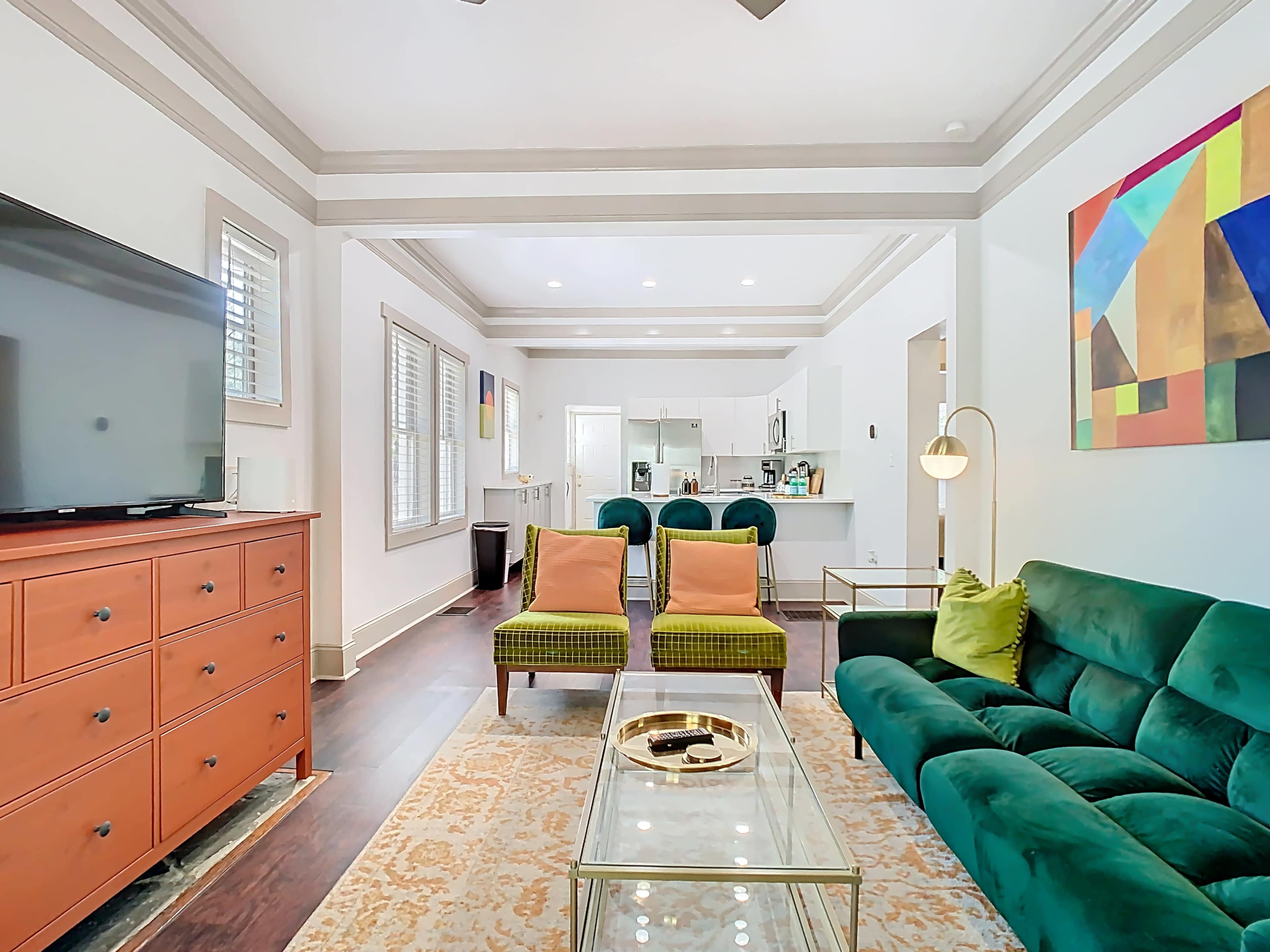 The image shows a modern living room featuring a green velvet sofa, two green and yellow accent chairs, a coffee table, and a television on a wooden dresser, with light-colored walls and artwork.