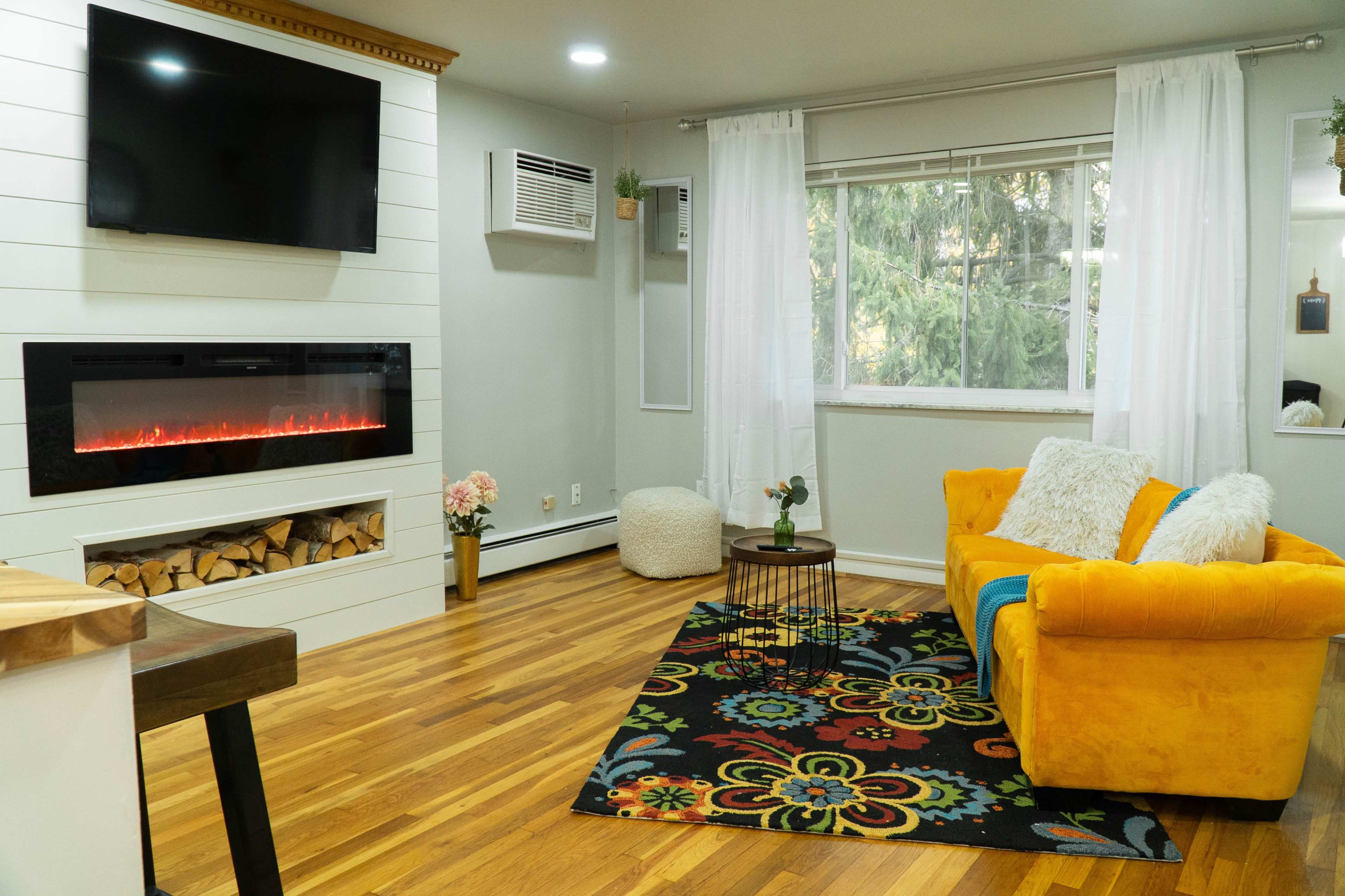 A cozy living room featuring a yellow armchair, a modern electric fireplace with a wood storage area, and a large window with sheer white curtains.
