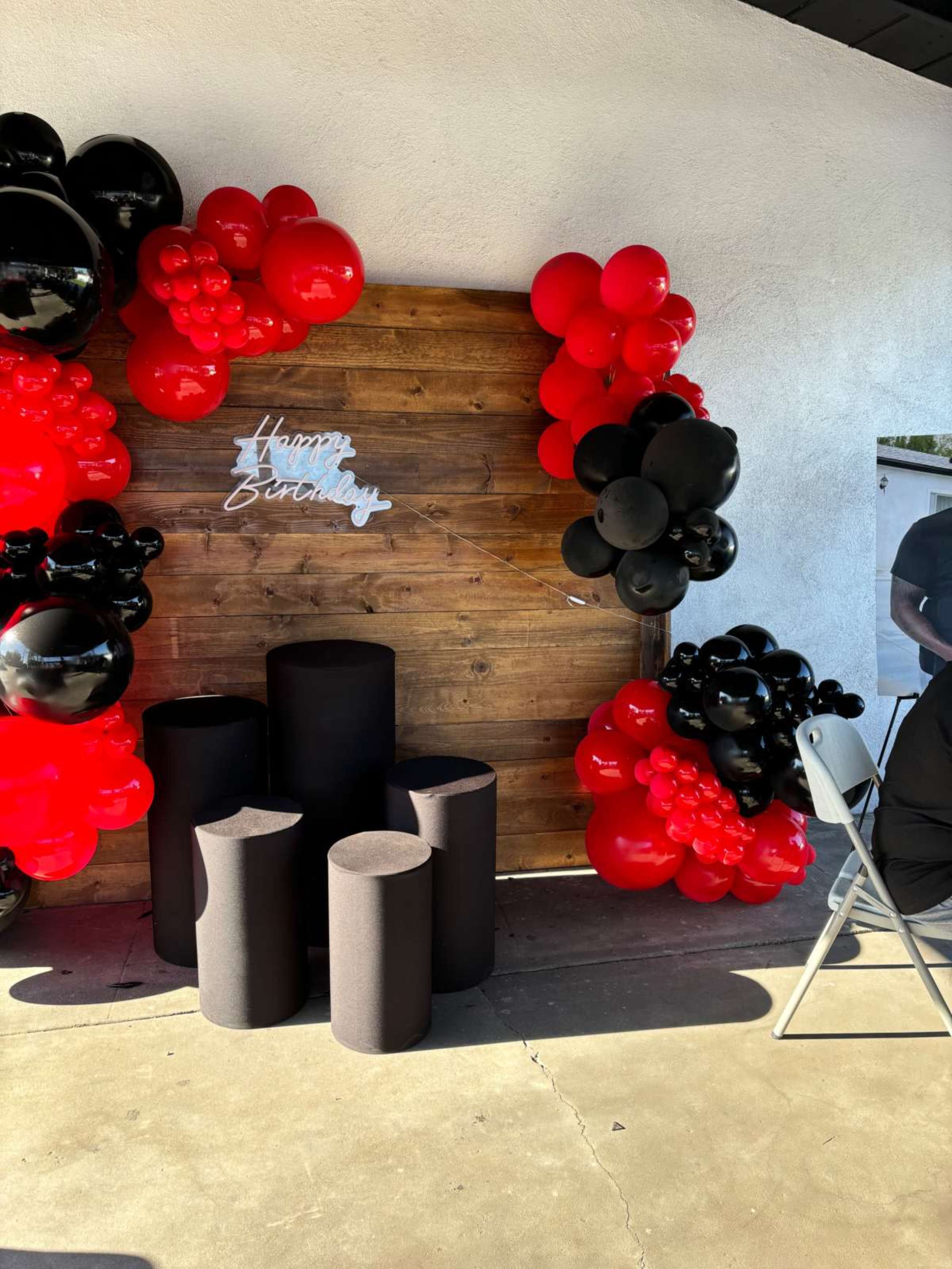 A birthday decoration setup featuring a wooden backdrop surrounded by red and black balloons, with three black cylindrical pedestals in front and a "Happy Birthday" sign.