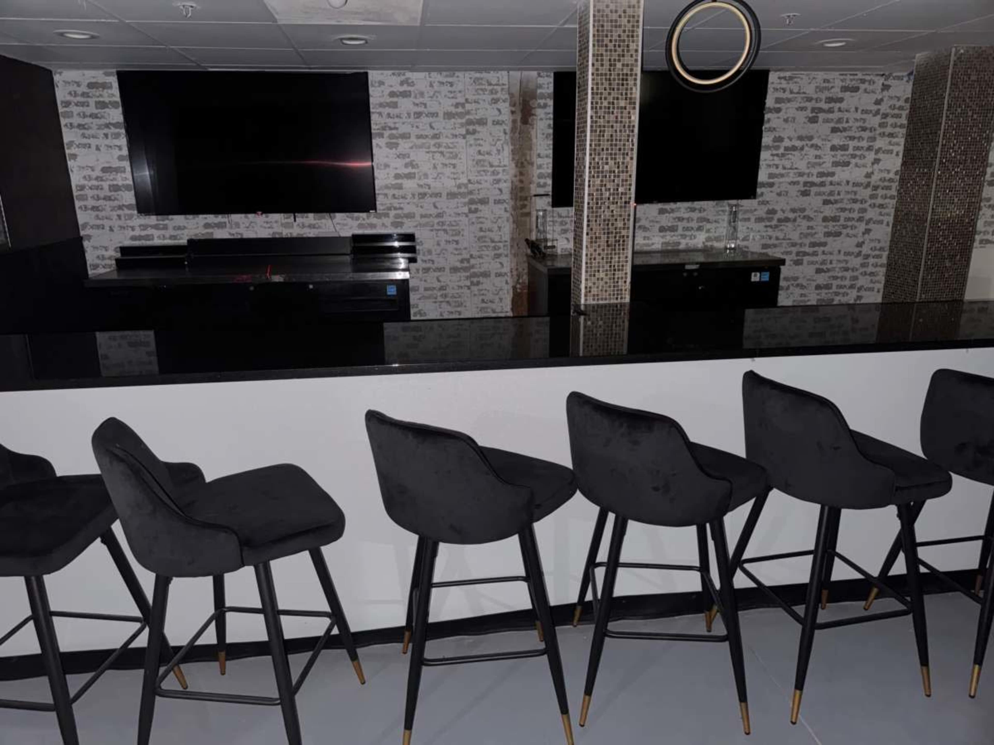 The image shows a modern bar area with several black upholstered stools lined up against a glossy black countertop, and two large screens mounted on a decorative wall in the background.