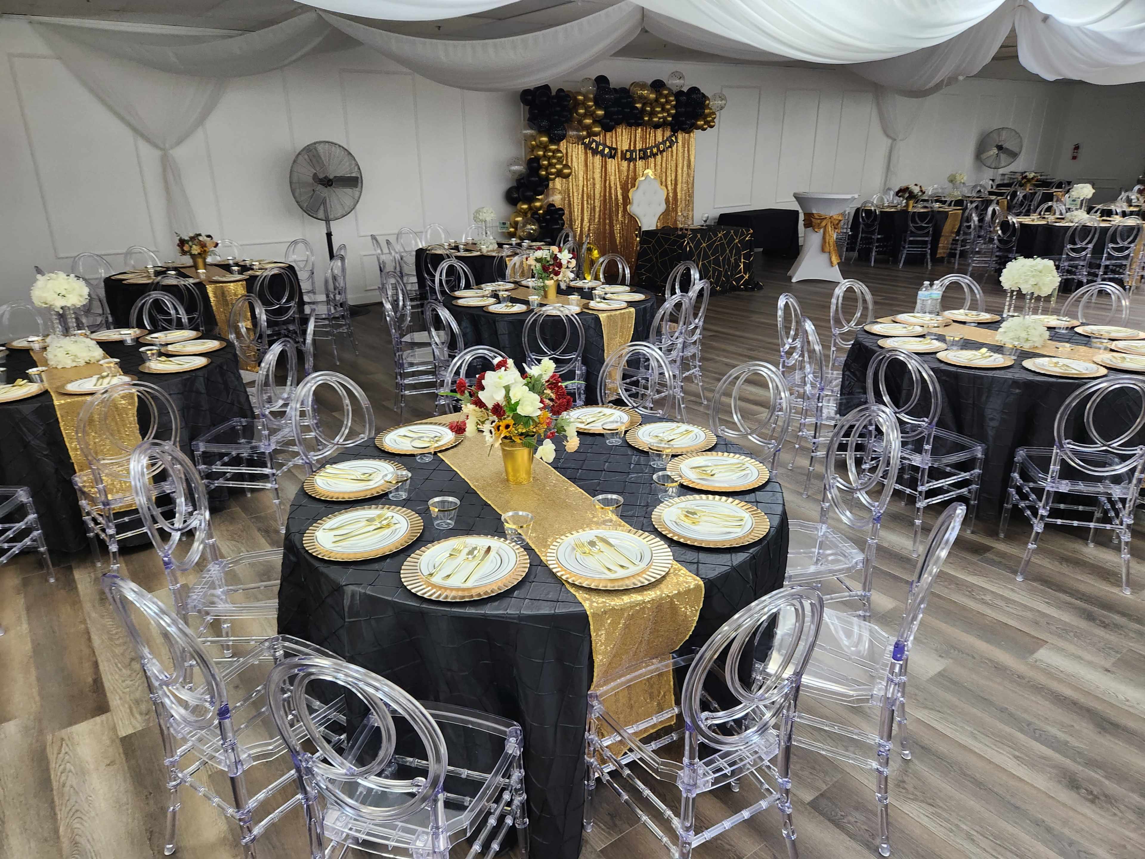 The room is set up for a formal event, featuring round tables covered with black tablecloths and adorned with gold decorations, clear acrylic chairs, and floral centerpieces.