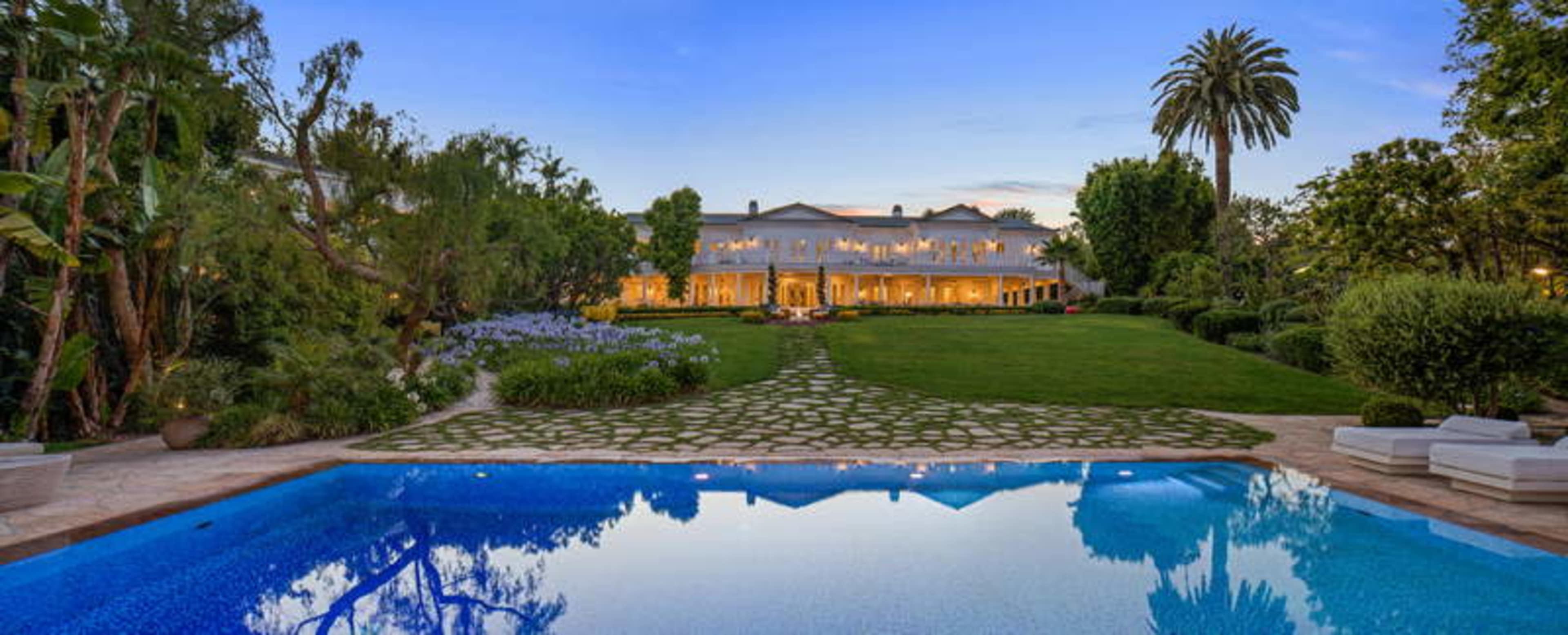 A large, luxurious mansion is reflected in a blue swimming pool, surrounded by well-maintained gardens and palm trees at dusk.