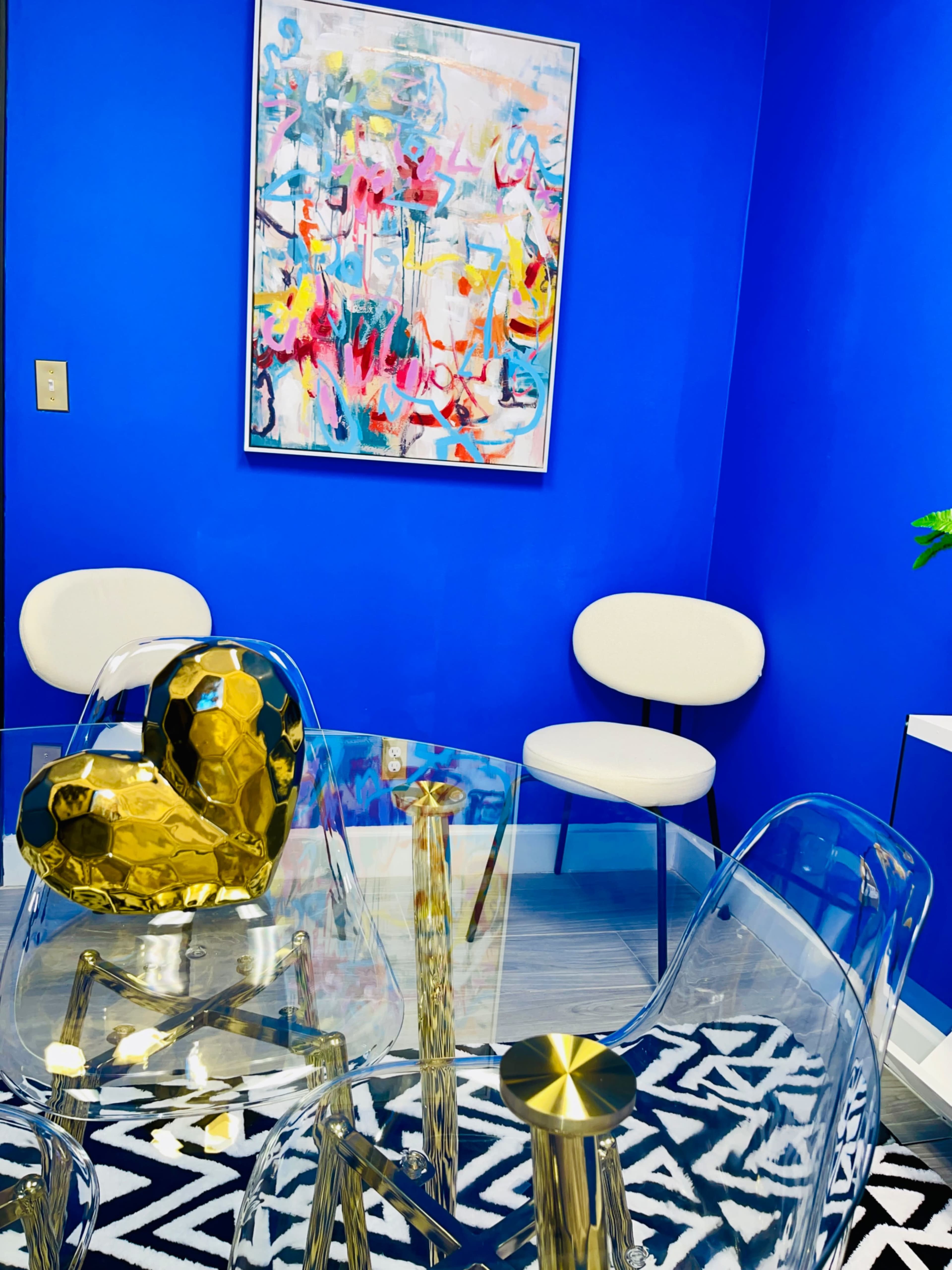 A modern seating area featuring a glass table, transparent chairs, and a blue wall adorned with an abstract painting.