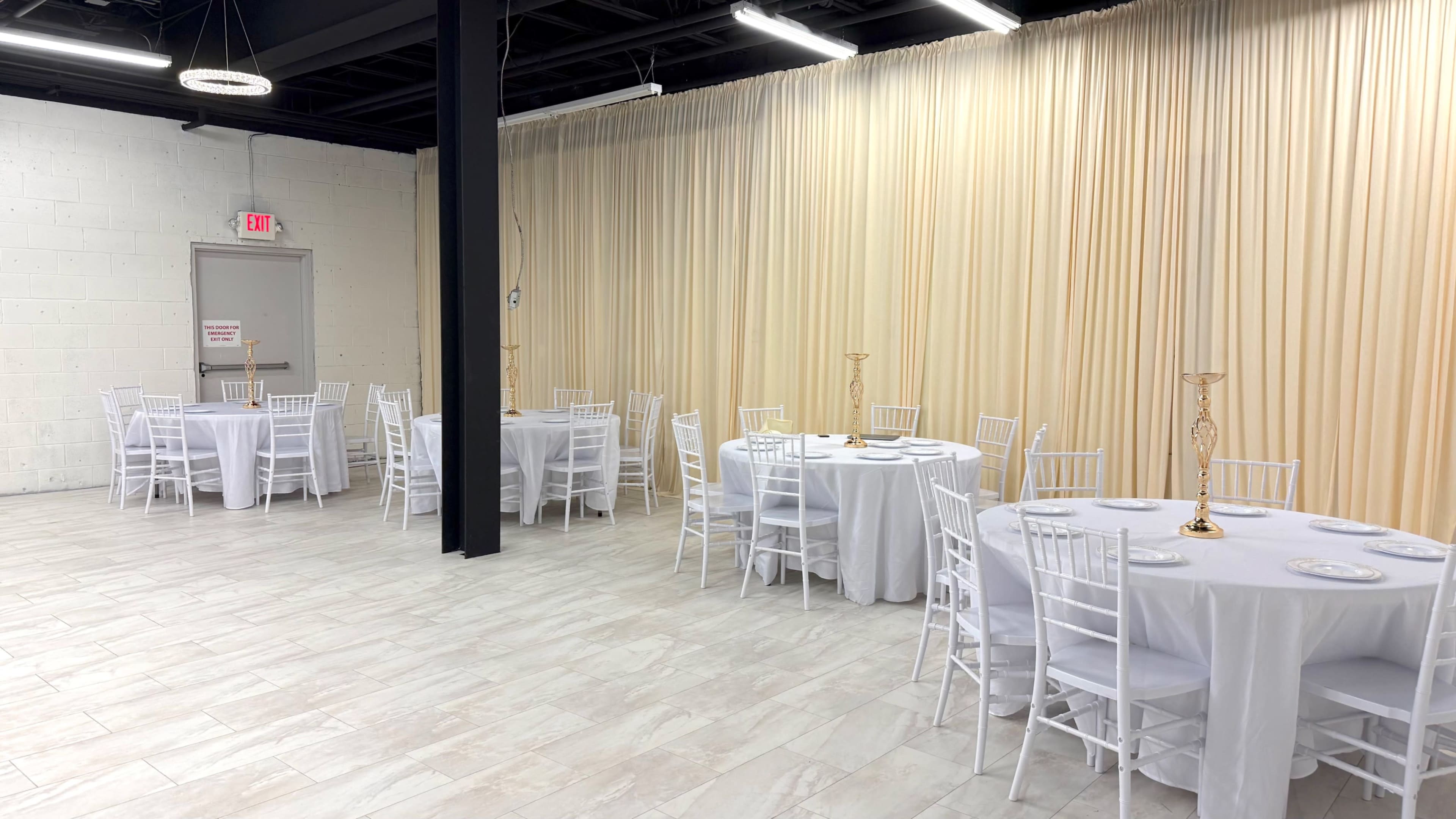 The image shows a spacious event room with several round tables covered in white tablecloths, surrounded by white Chiavari chairs, and a backdrop of beige curtains.