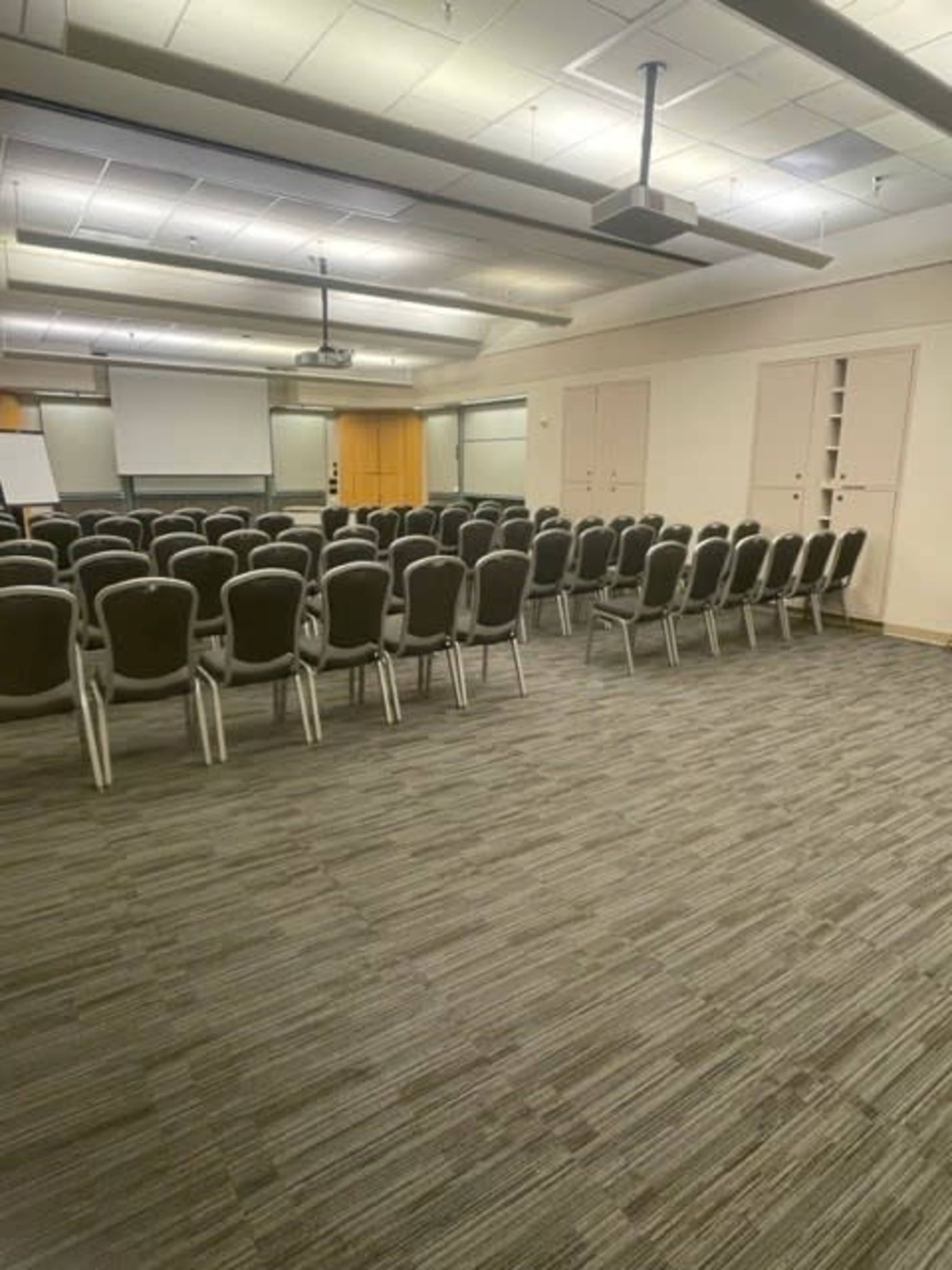 A spacious conference room arranged with rows of chairs facing a blank presentation screen and a projector.