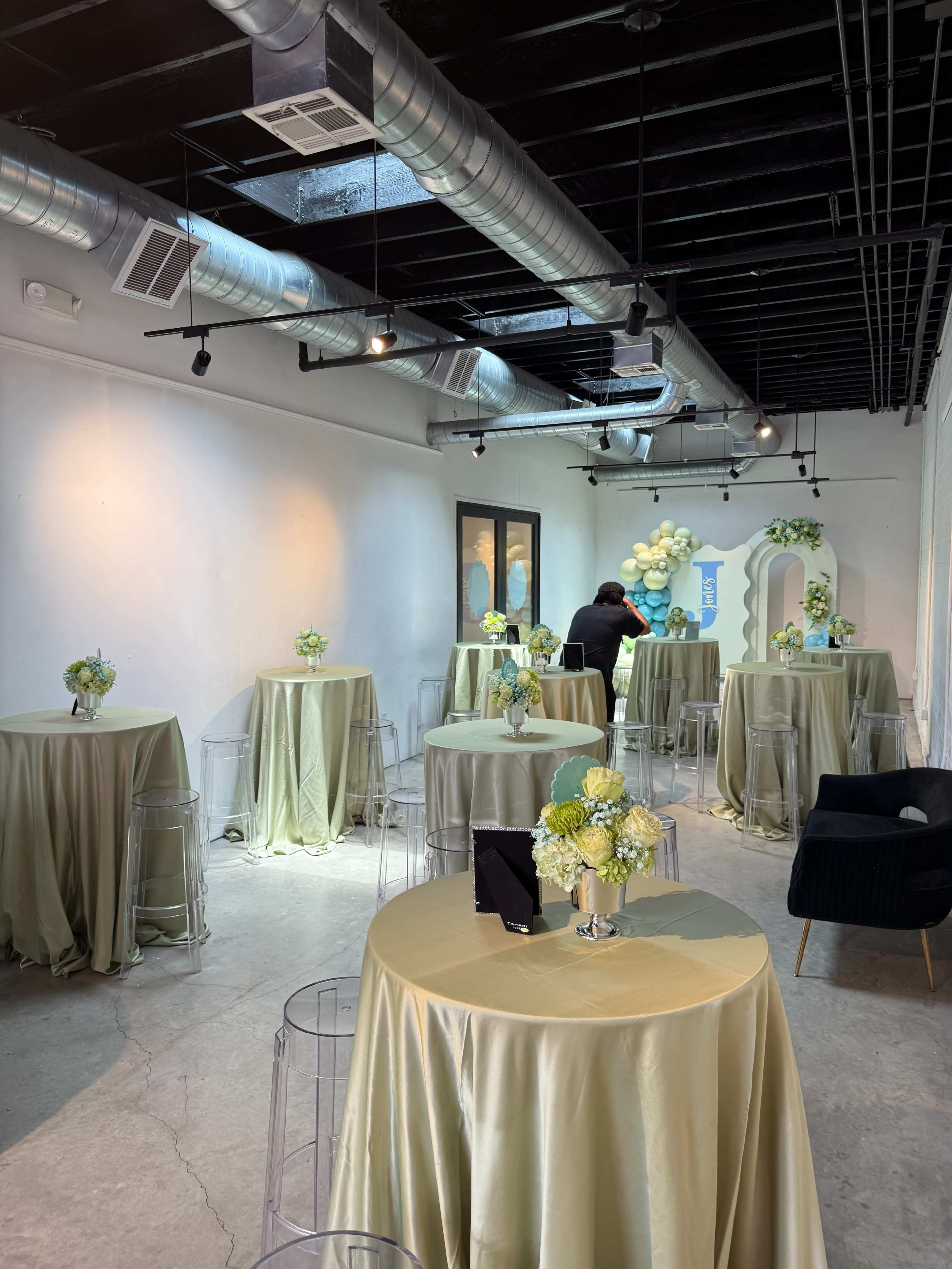The image shows a decorated event space with round tables covered in light-colored tablecloths and floral centerpieces, featuring a person arranging decor in the background.