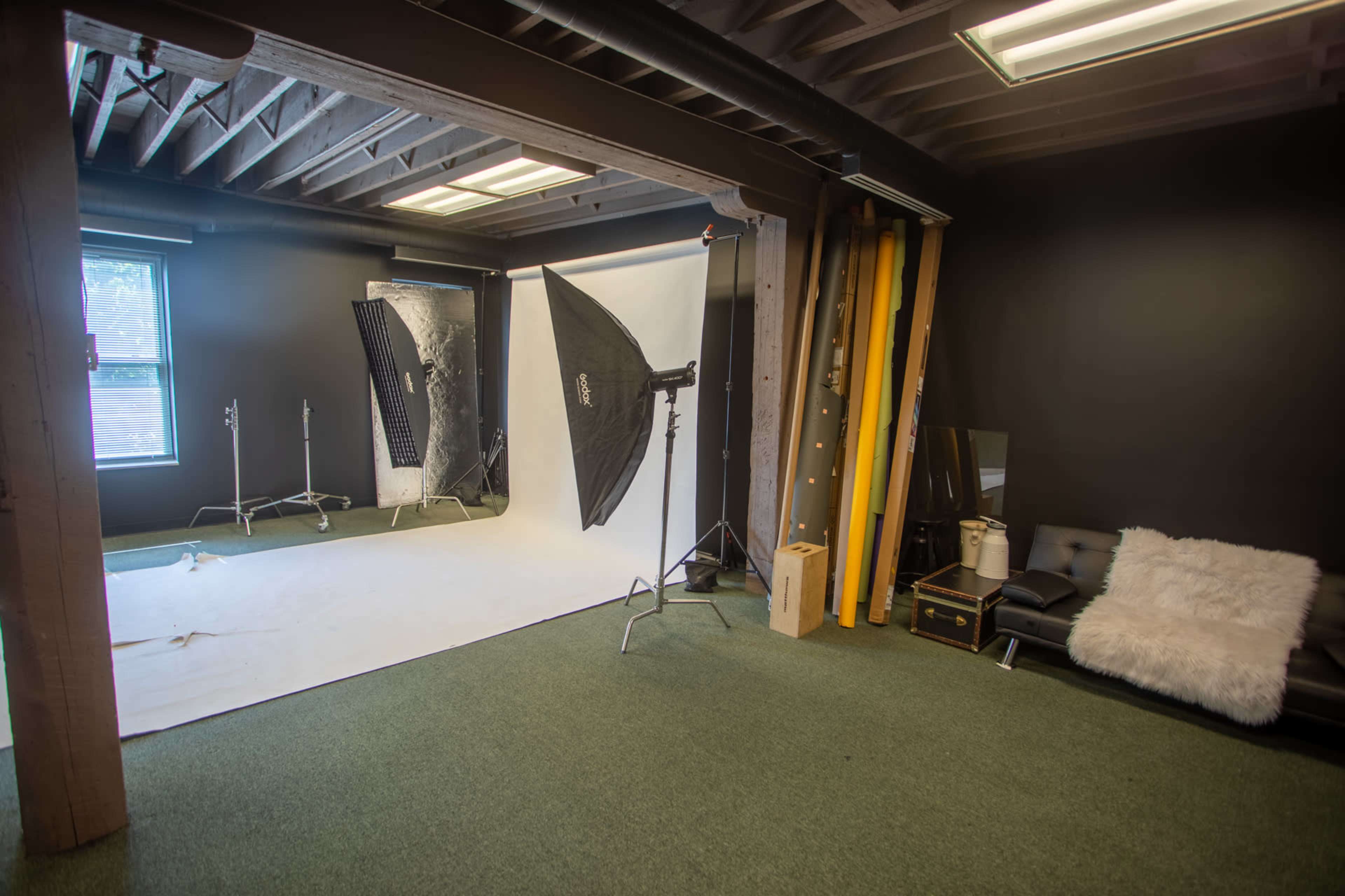 A photography studio featuring a white backdrop, two softbox lights on stands, and a dark carpeted area with a black couch and additional props in the corner.