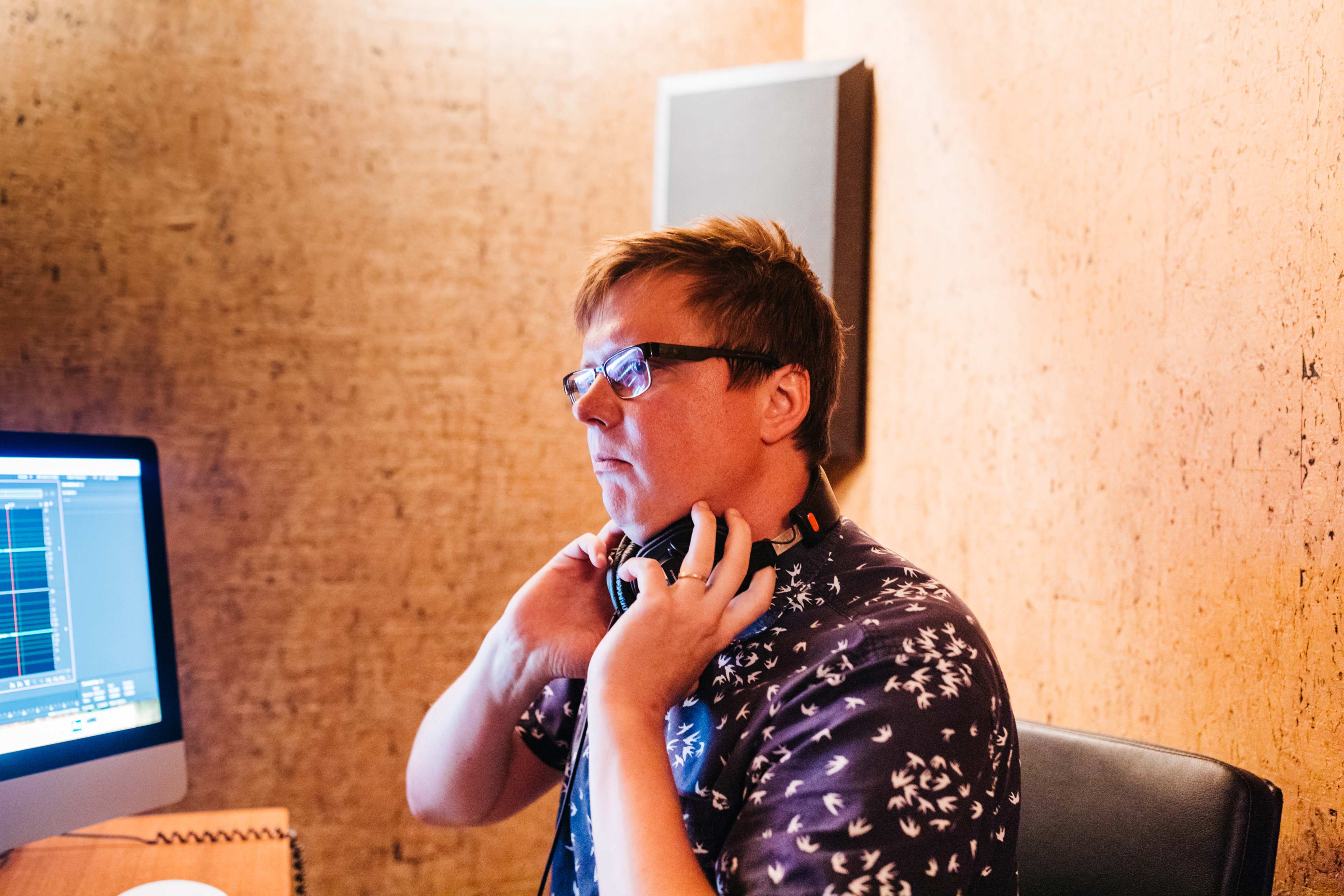 A person adjusts headphones while sitting in front of a computer in a room with soundproofing.