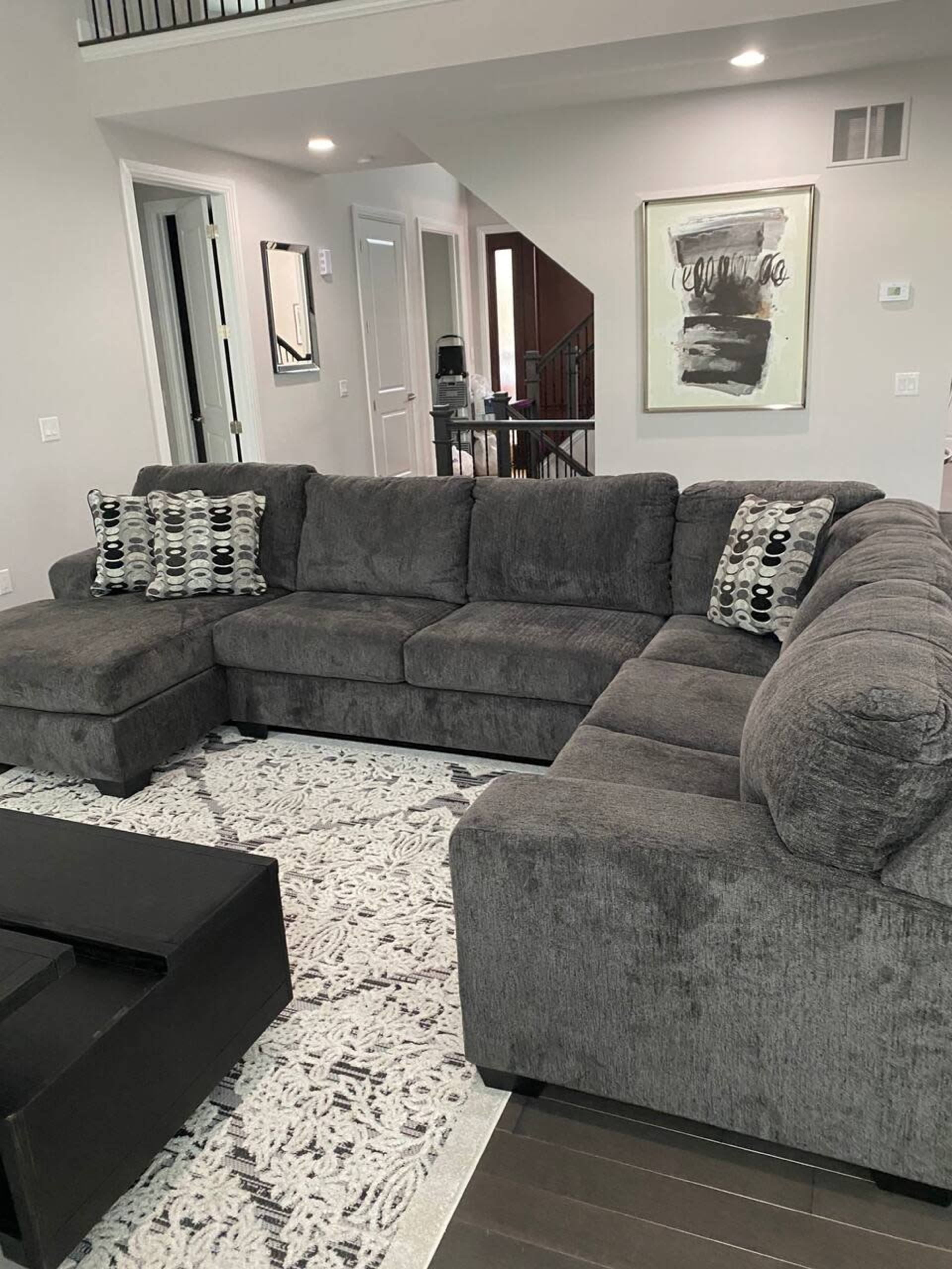 A gray sectional sofa is positioned in a living room with light walls, a patterned area rug, and a large piece of abstract art on the wall.
