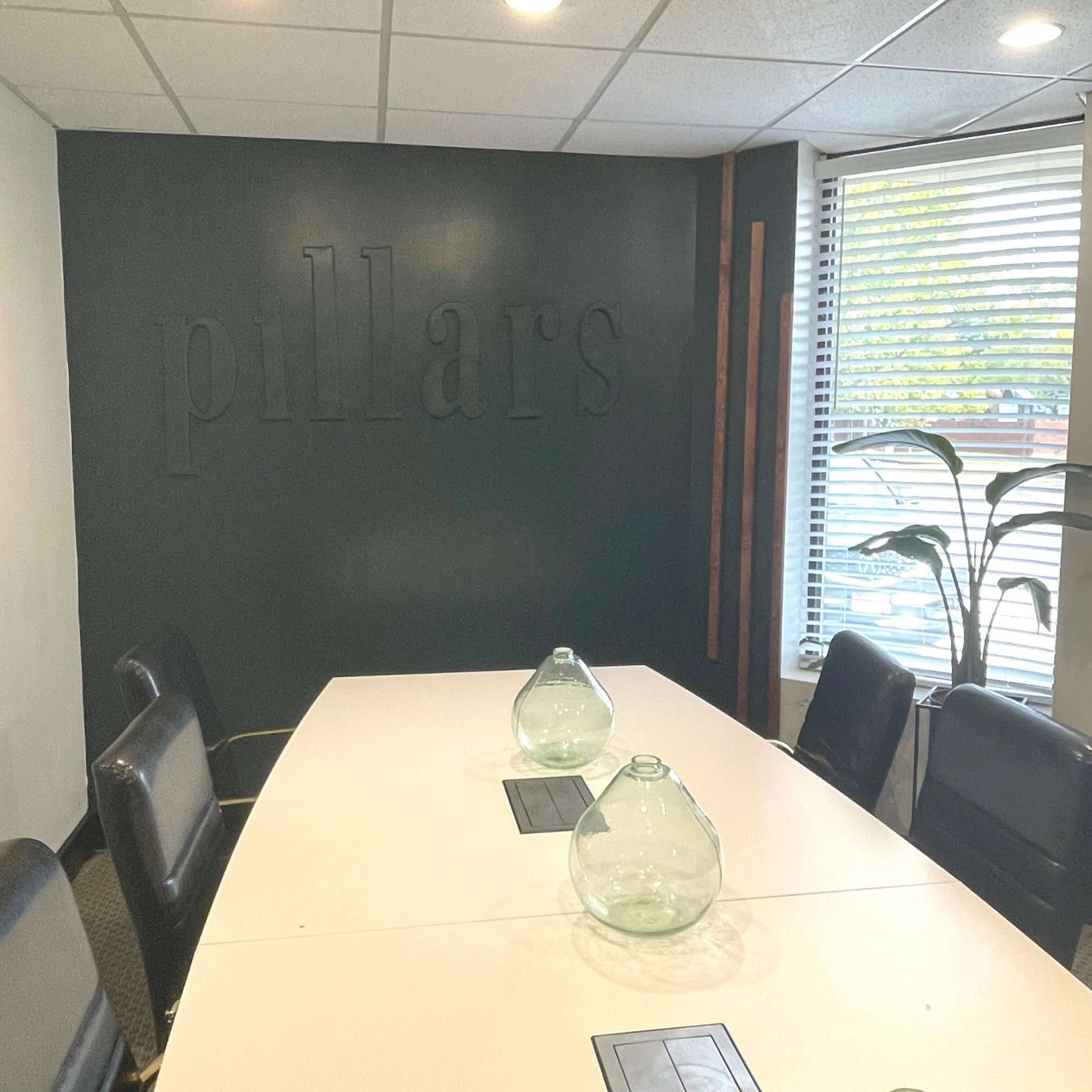 A conference room features a long white table, two glass vases, black chairs, and a green wall with the word "pillars" carved into it.