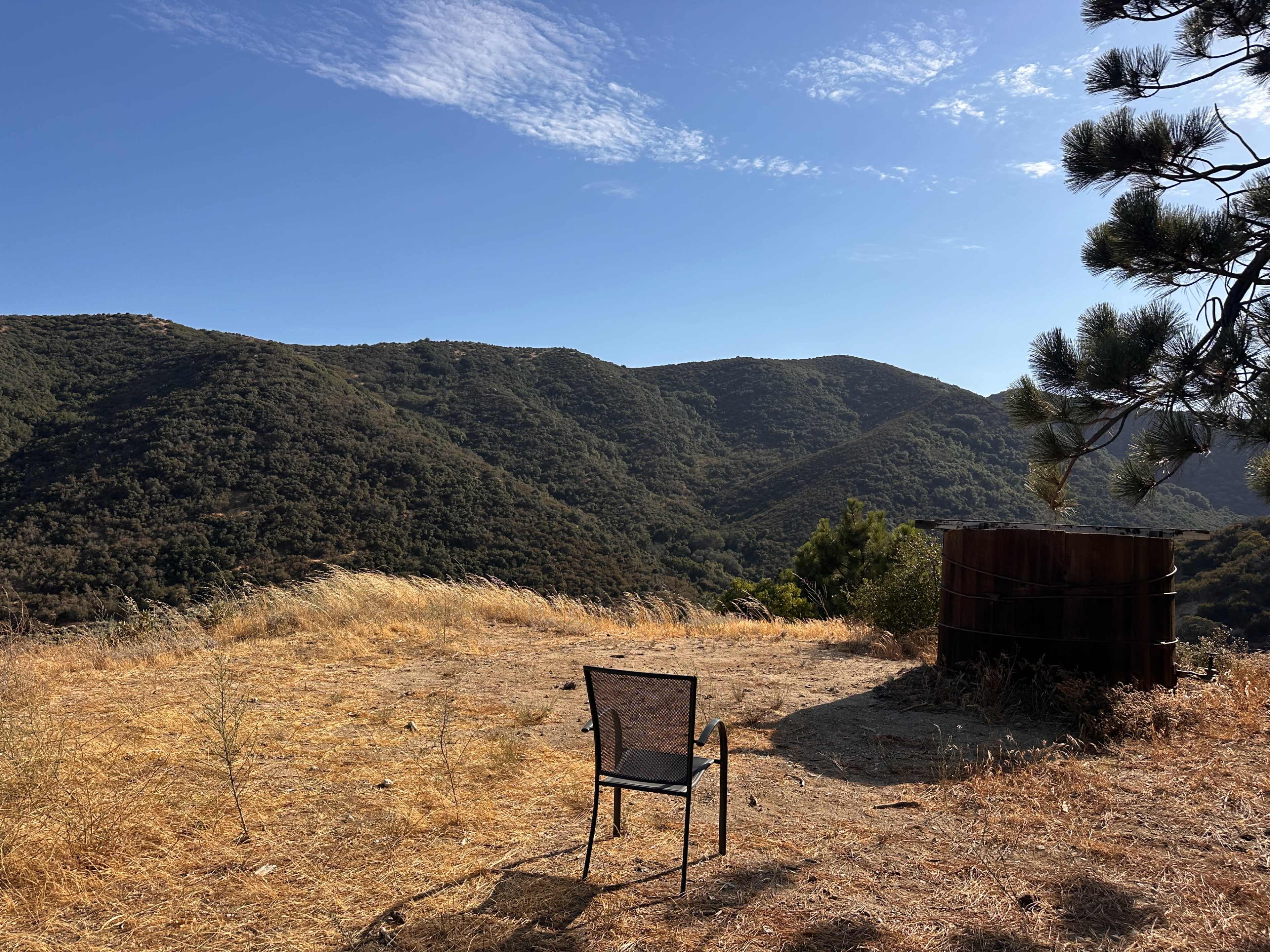 A metal chair sits on dry grass overlooking a mountainous landscape under a clear blue sky.