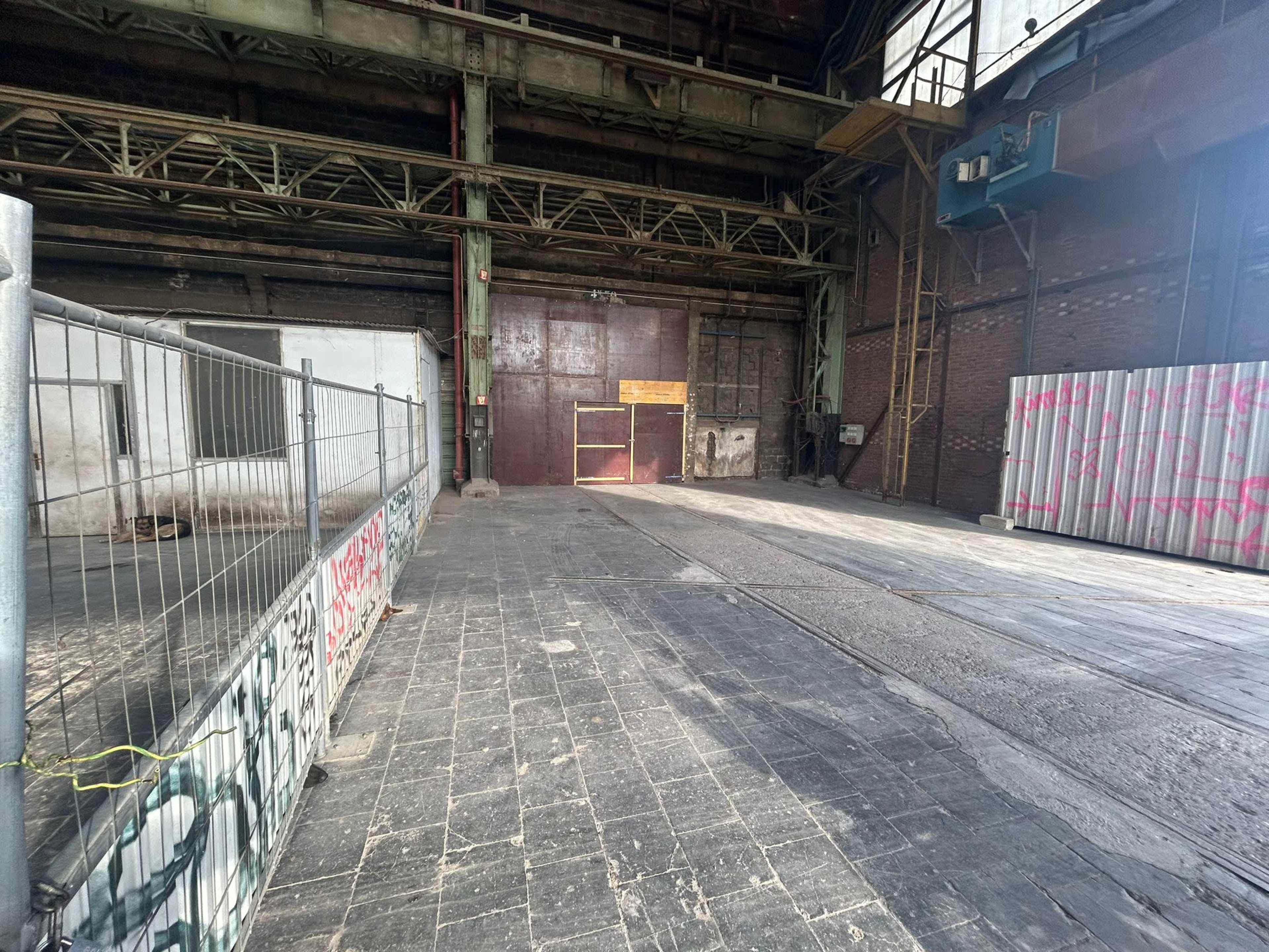 A large, empty industrial space with a gravel floor, metal beams, and a partially fenced-off area.