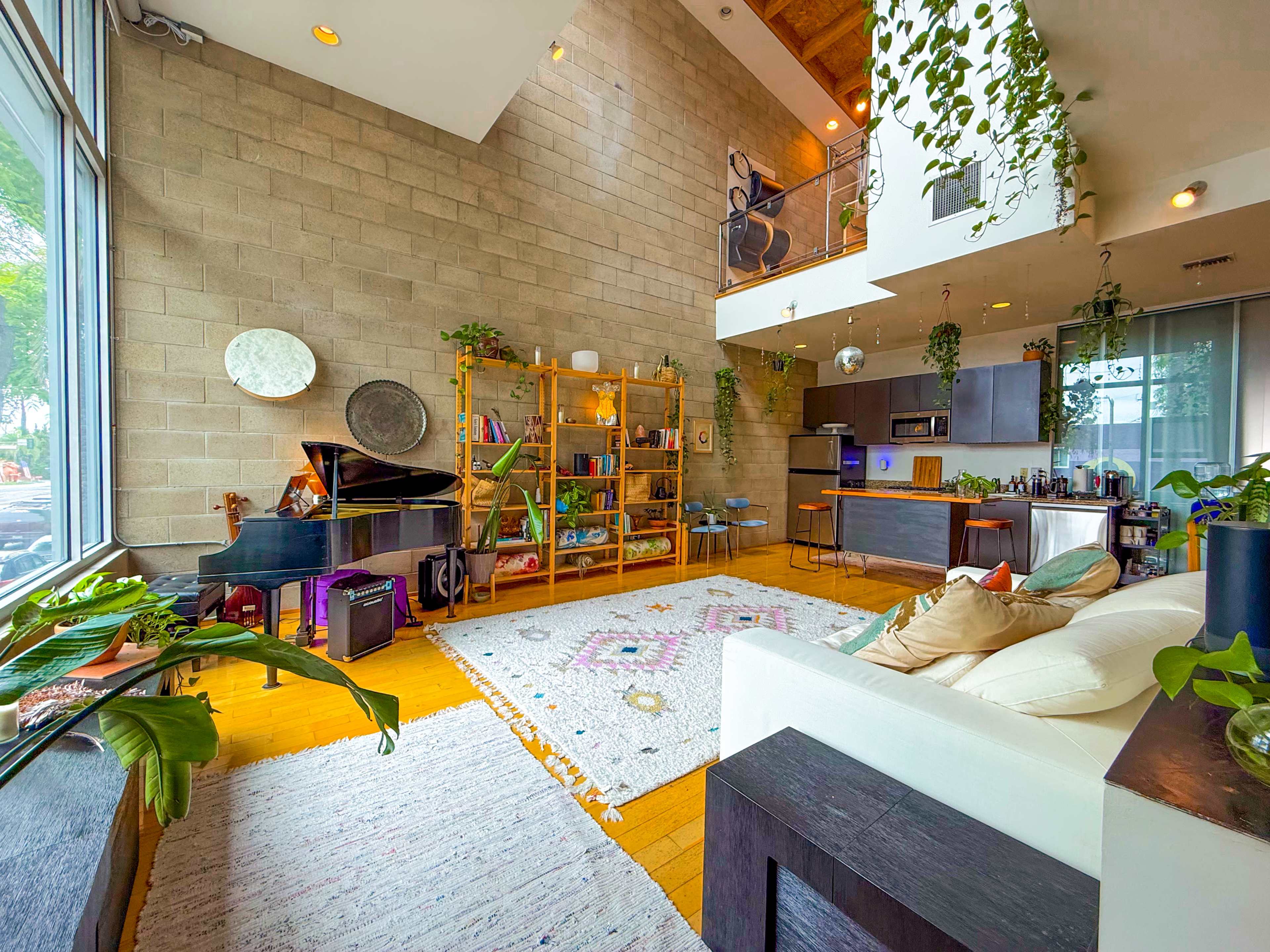 The image depicts an open-concept living space with a high ceiling, featuring a piano, a bookshelf, and a cozy seating area centered on a patterned rug.
