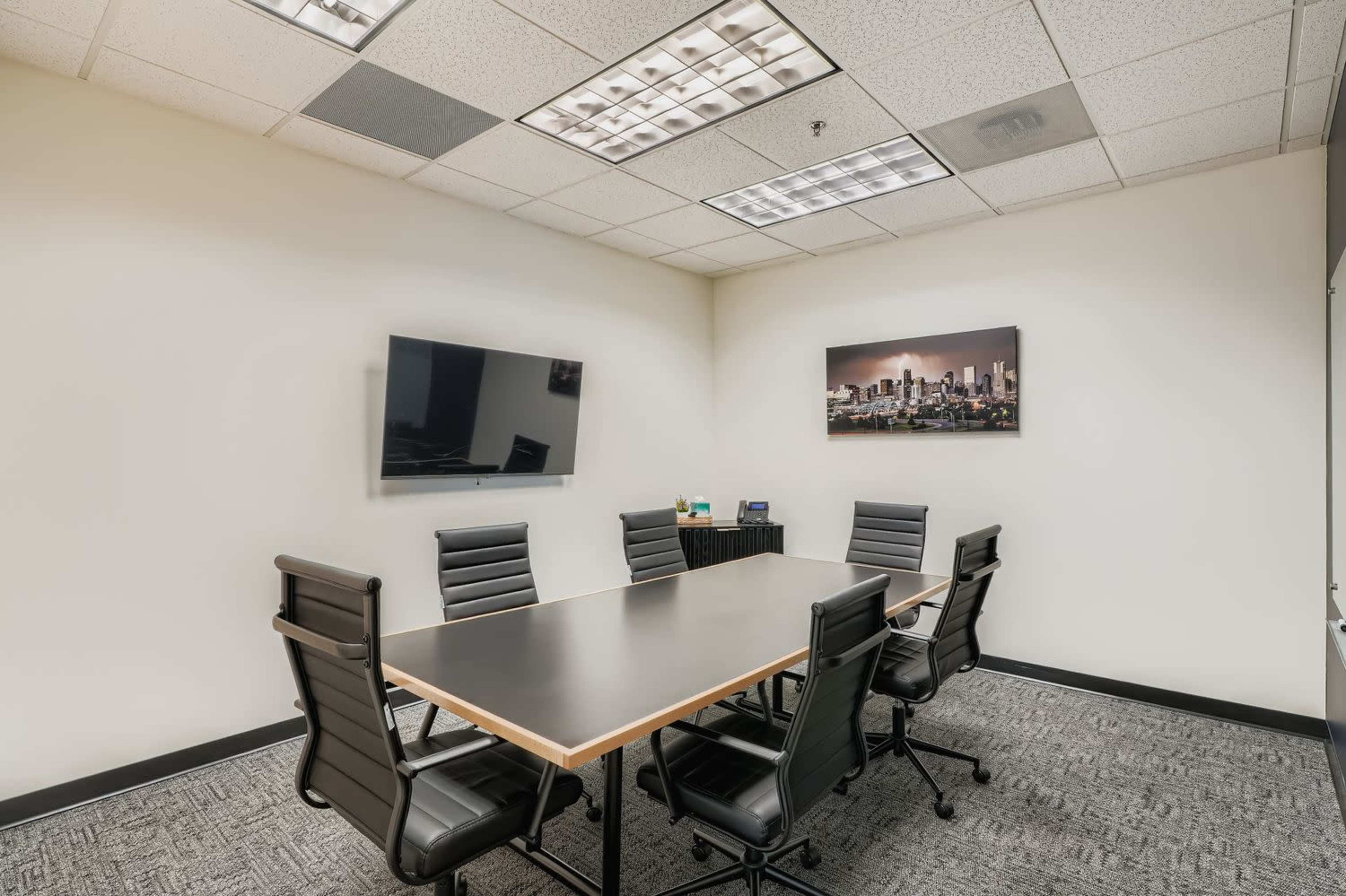 The image shows a modern conference room with a large table surrounded by black chairs, a wall-mounted television, and a cityscape photograph.