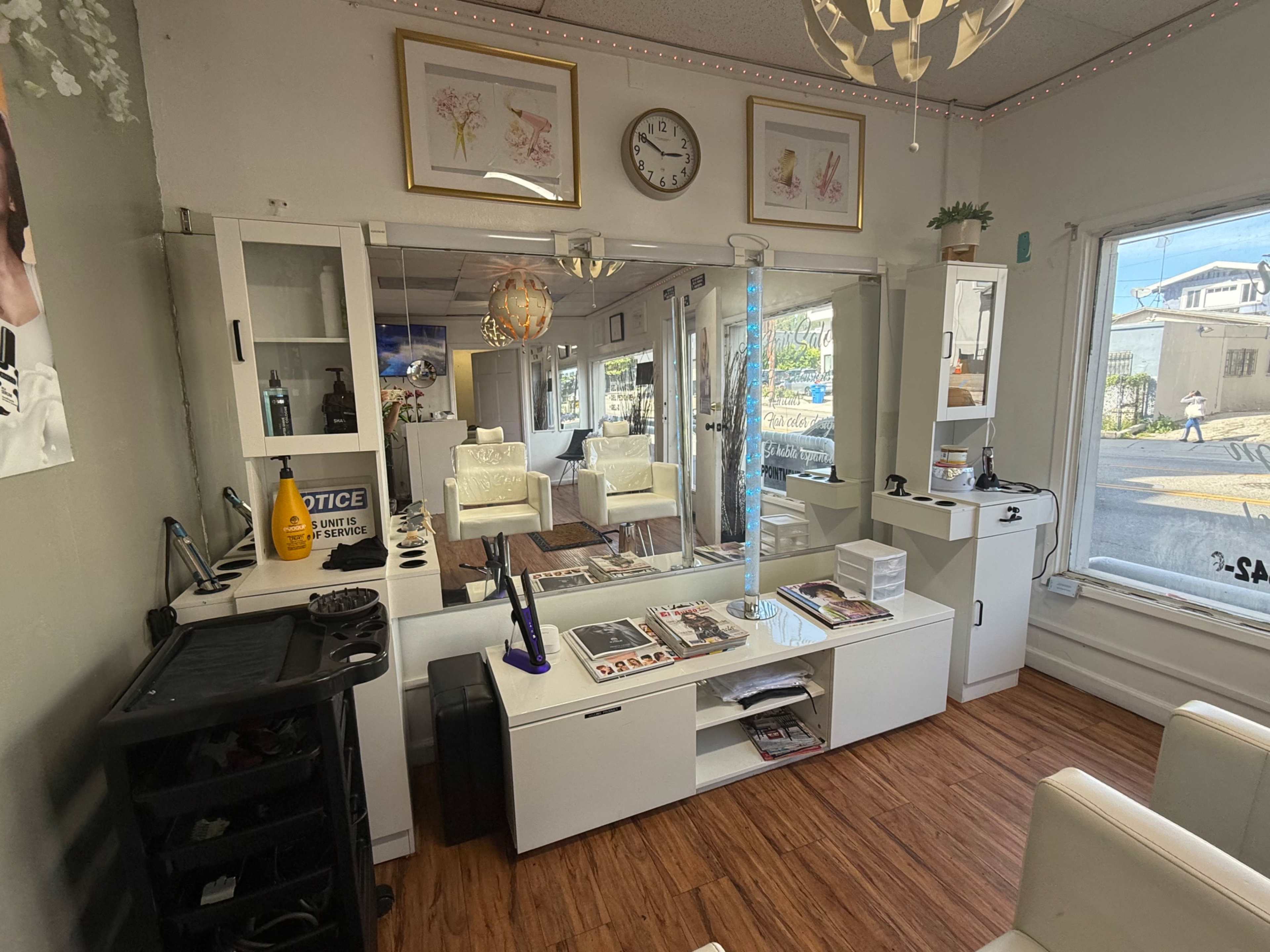 The image shows the interior of a salon with a reception area featuring a white counter, a mirror, and various chairs, all illuminated by natural light through large windows.