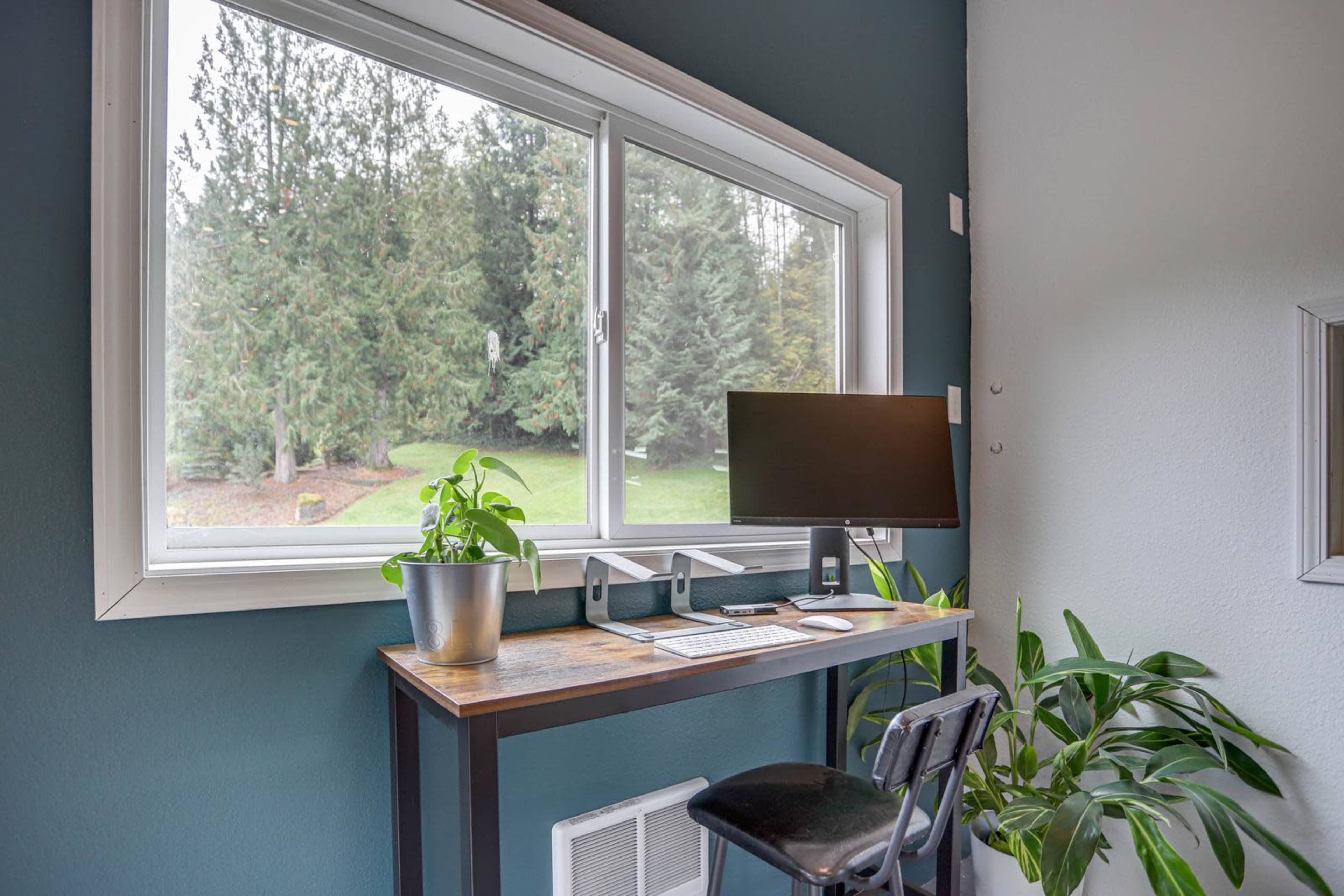 A modern workspace with a computer monitor, keyboard, and a potted plant, situated by a large window overlooking a green landscape.