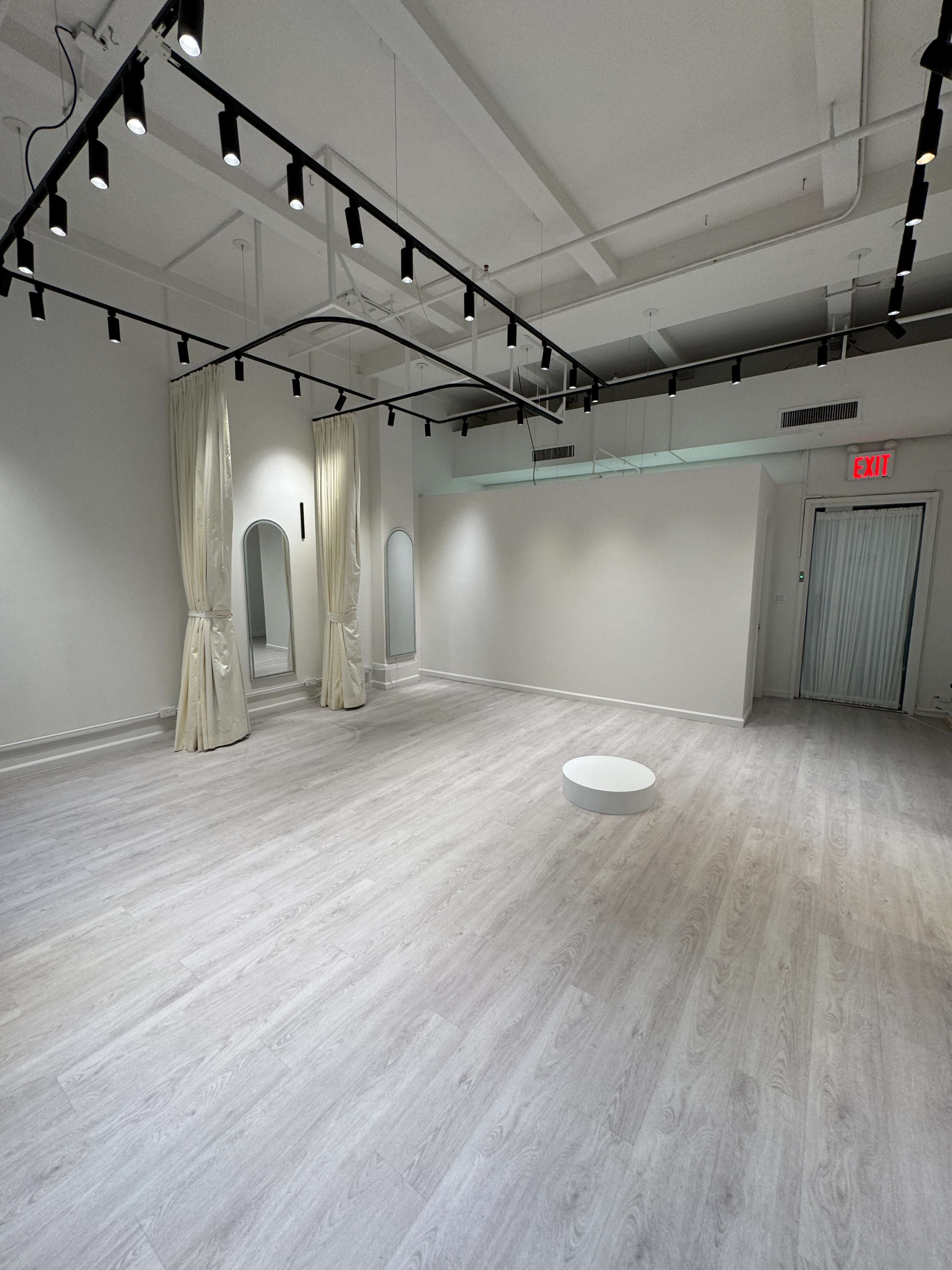 The image shows a spacious, minimalistic indoor room with light-colored wooden flooring, equipped with wall-mounted lights and a few decorative mirrors.