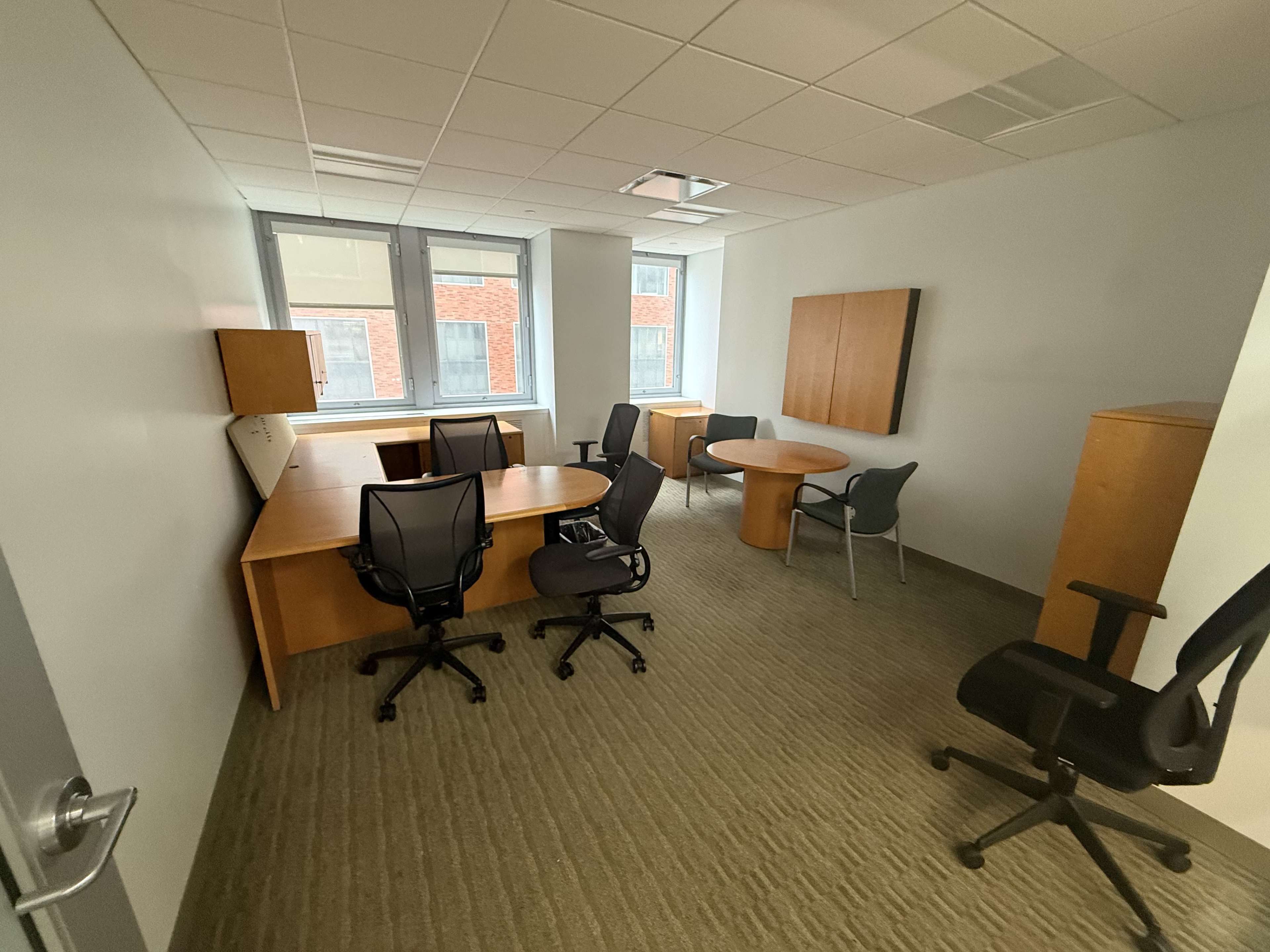 The image shows a sparsely furnished office space with two large desks, several office chairs, and two windows allowing natural light.