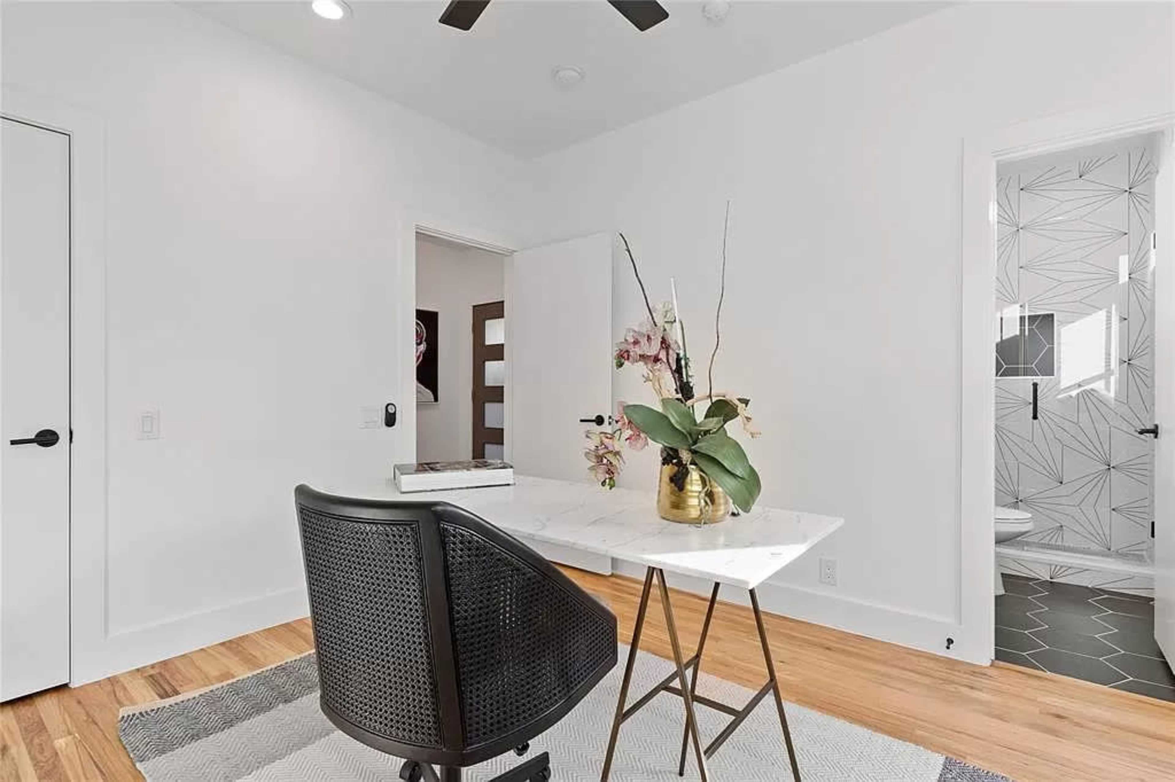 A modern workspace features a marble-topped desk with a plant arrangement, a black chair, and a doorway leading to a bathroom with geometric tile.