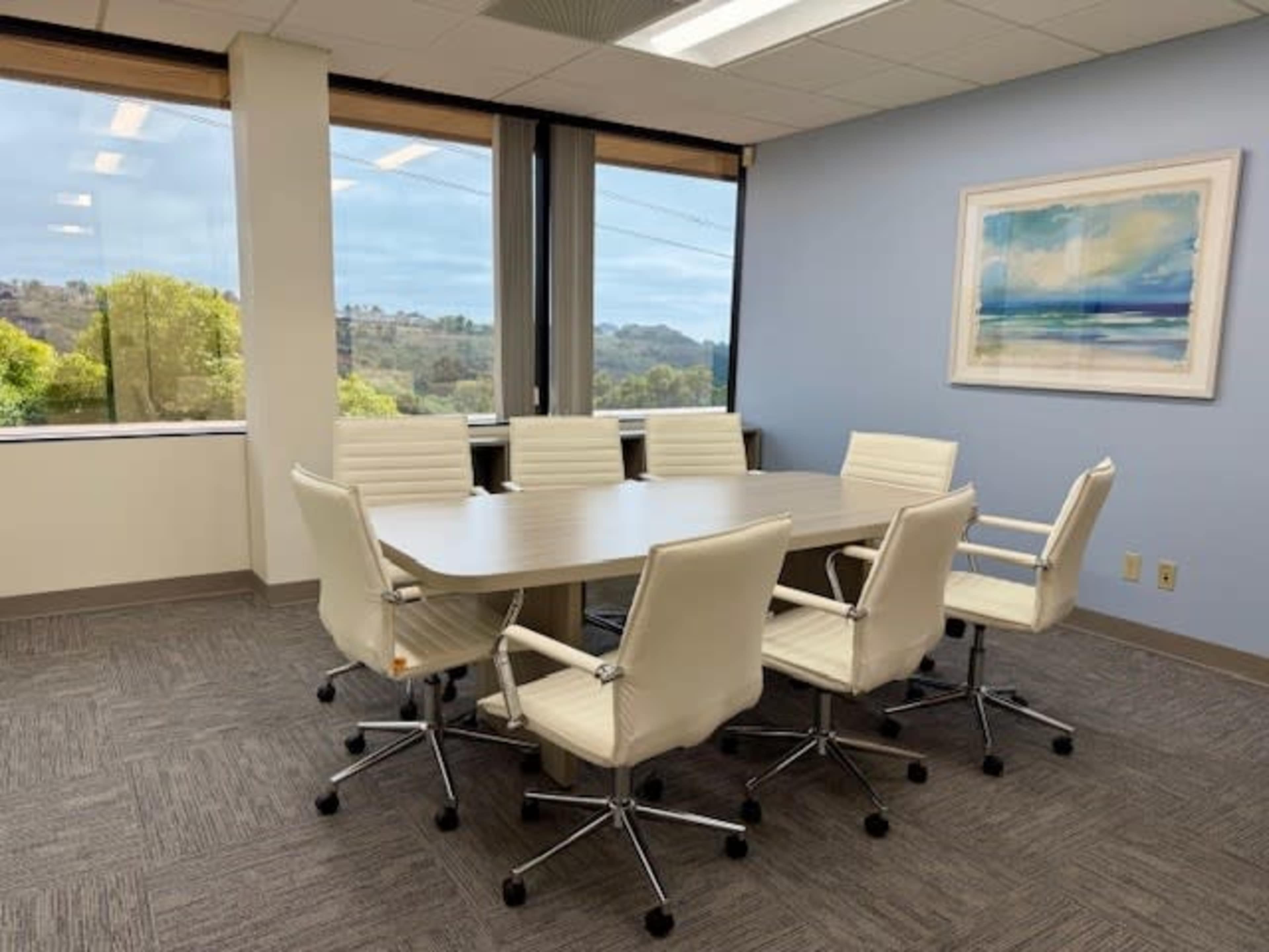 A conference room features a large wooden table surrounded by eight white swivel chairs, with large windows showcasing a view of greenery outside.