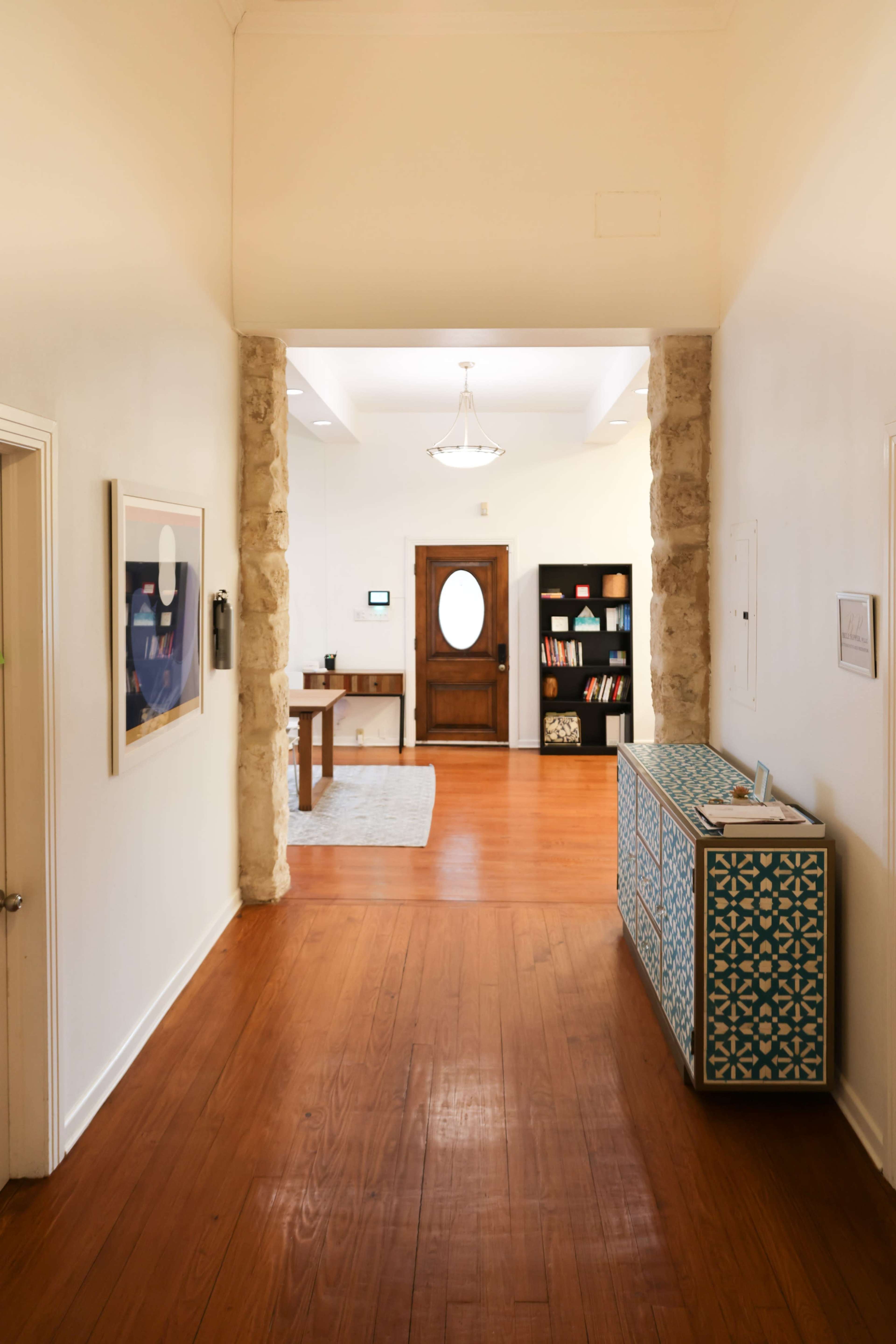 A brightly lit hallway features wooden flooring, framed artwork on the walls, and a patterned console table, leading to a door with an oval window and a room beyond.