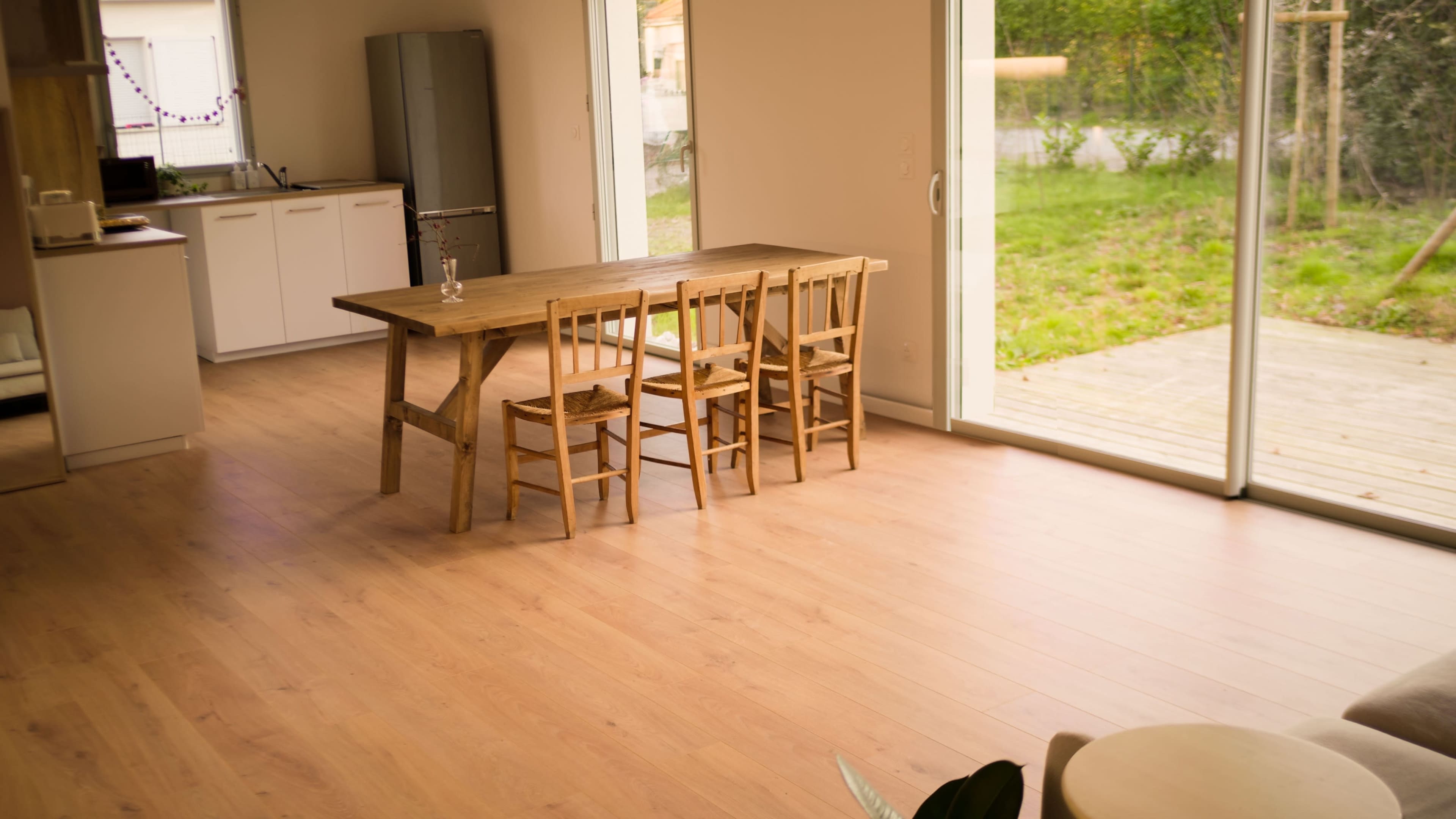 A wooden dining table with four chairs is positioned in a bright room that opens to a garden.