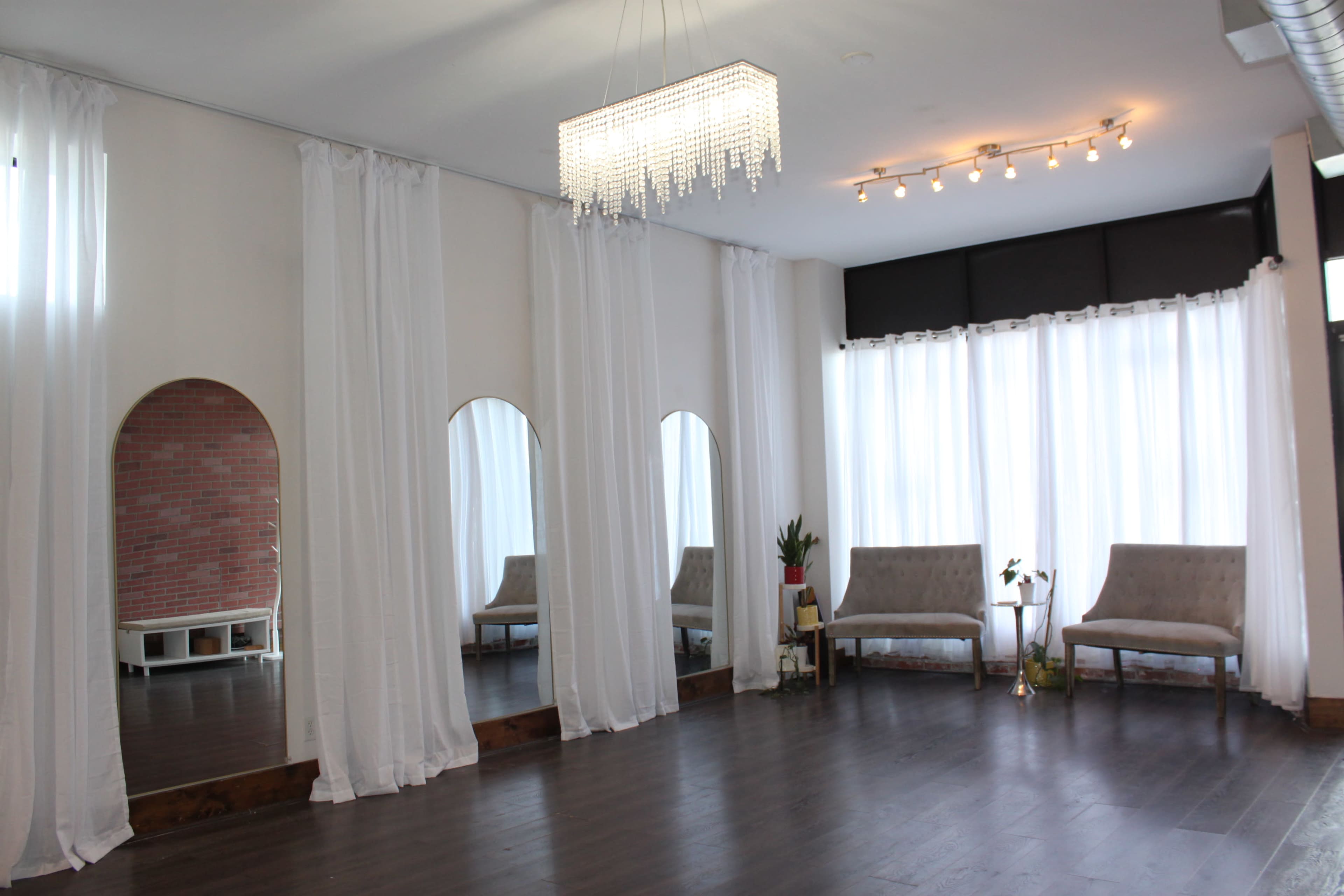 The image shows a bright, spacious room with white drapes, two large mirrors, minimalist seating, and a chandelier hanging from the ceiling.
