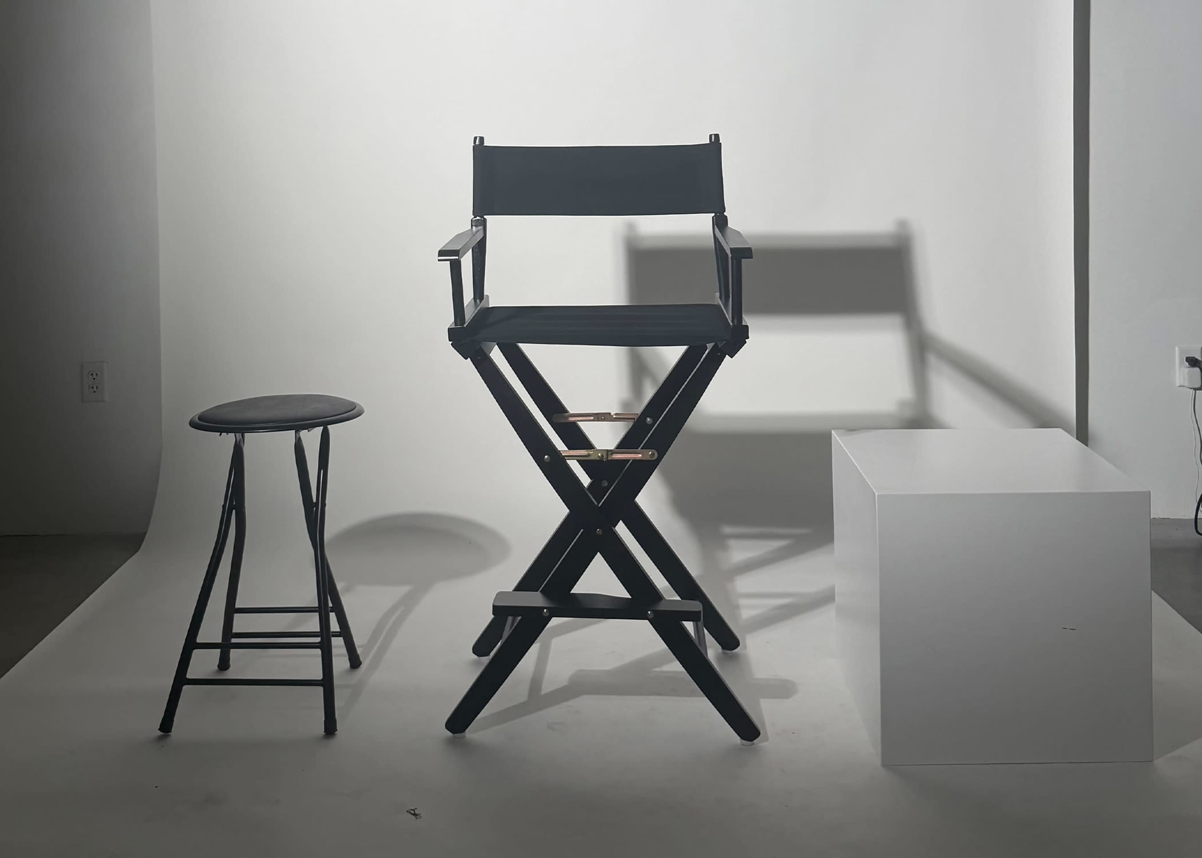 The image features a director's chair, a stool, and a white cube positioned against a neutral backdrop.