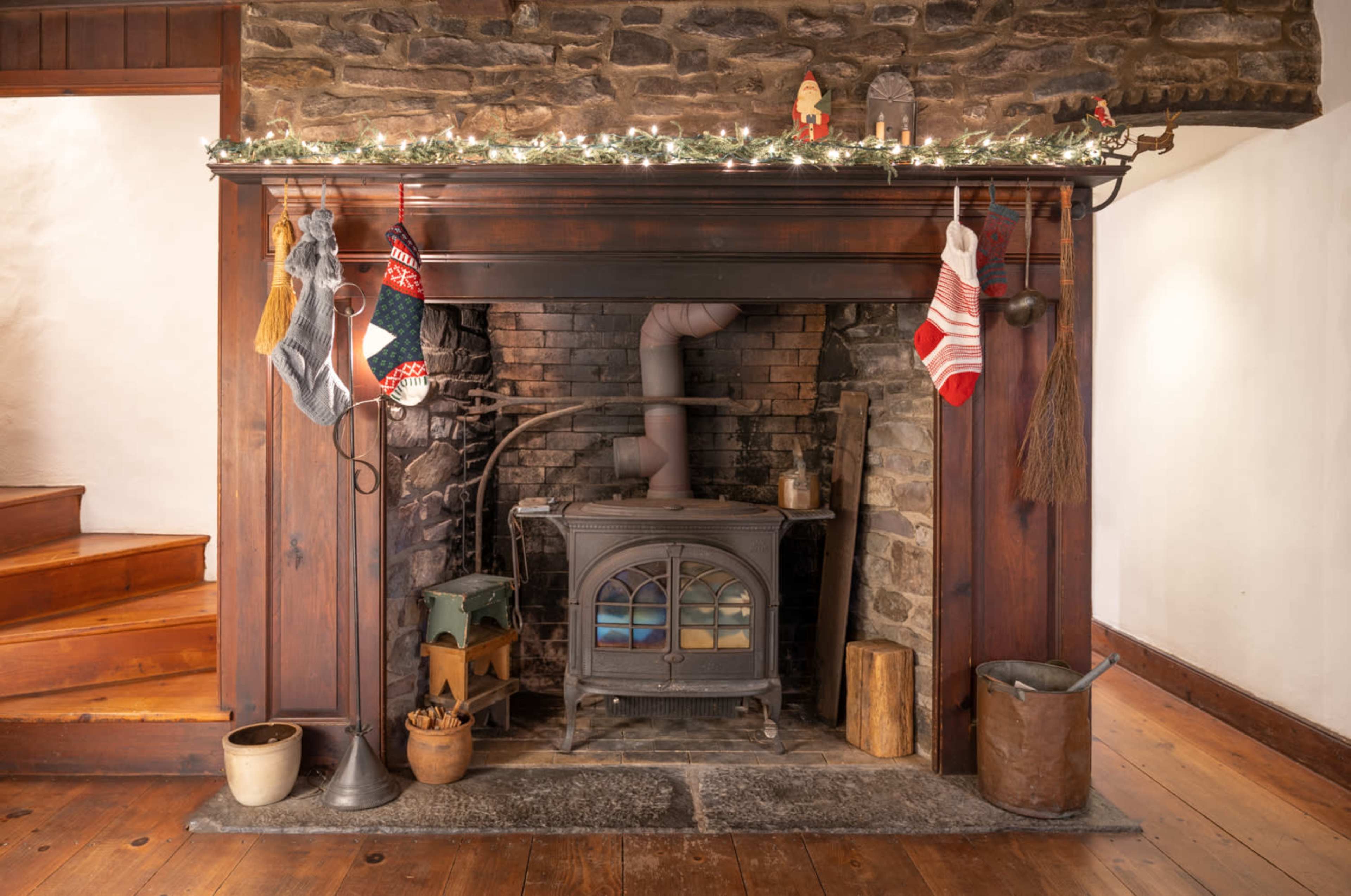 A stone fireplace is adorned with stockings and festive decorations, featuring a wood stove and various tools nearby.