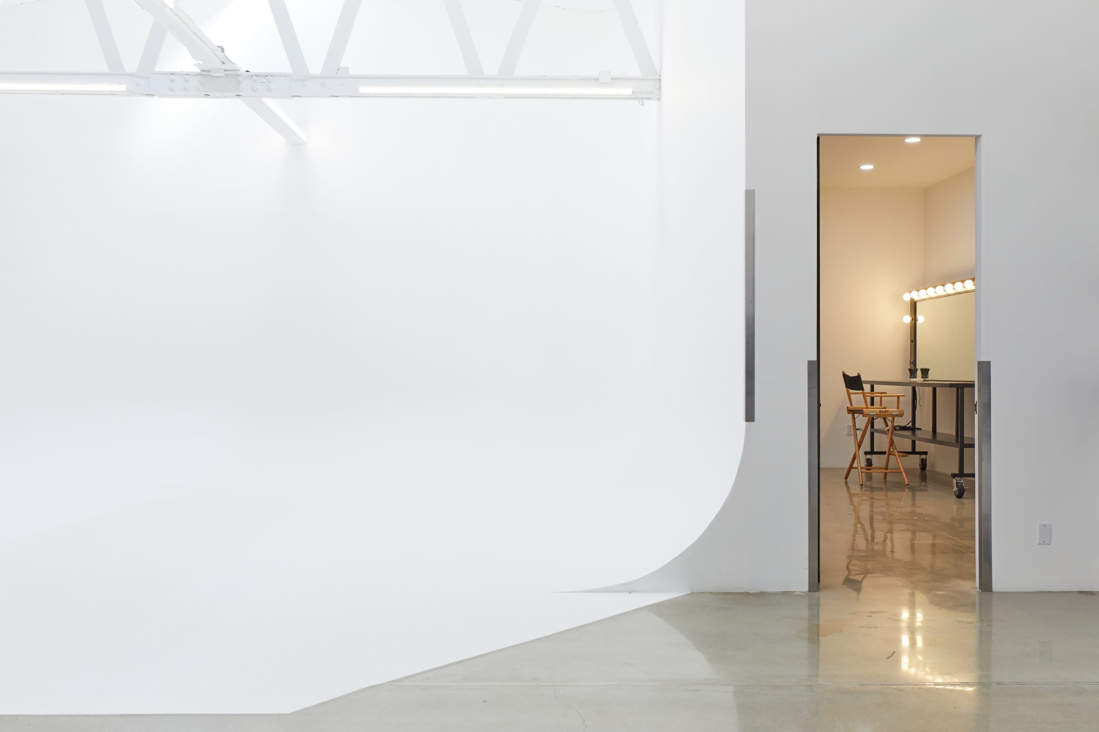 The image shows a minimalistic, white studio space with a seamless backdrop and an open doorway leading to a small, illuminated dressing area with a mirror and a chair.