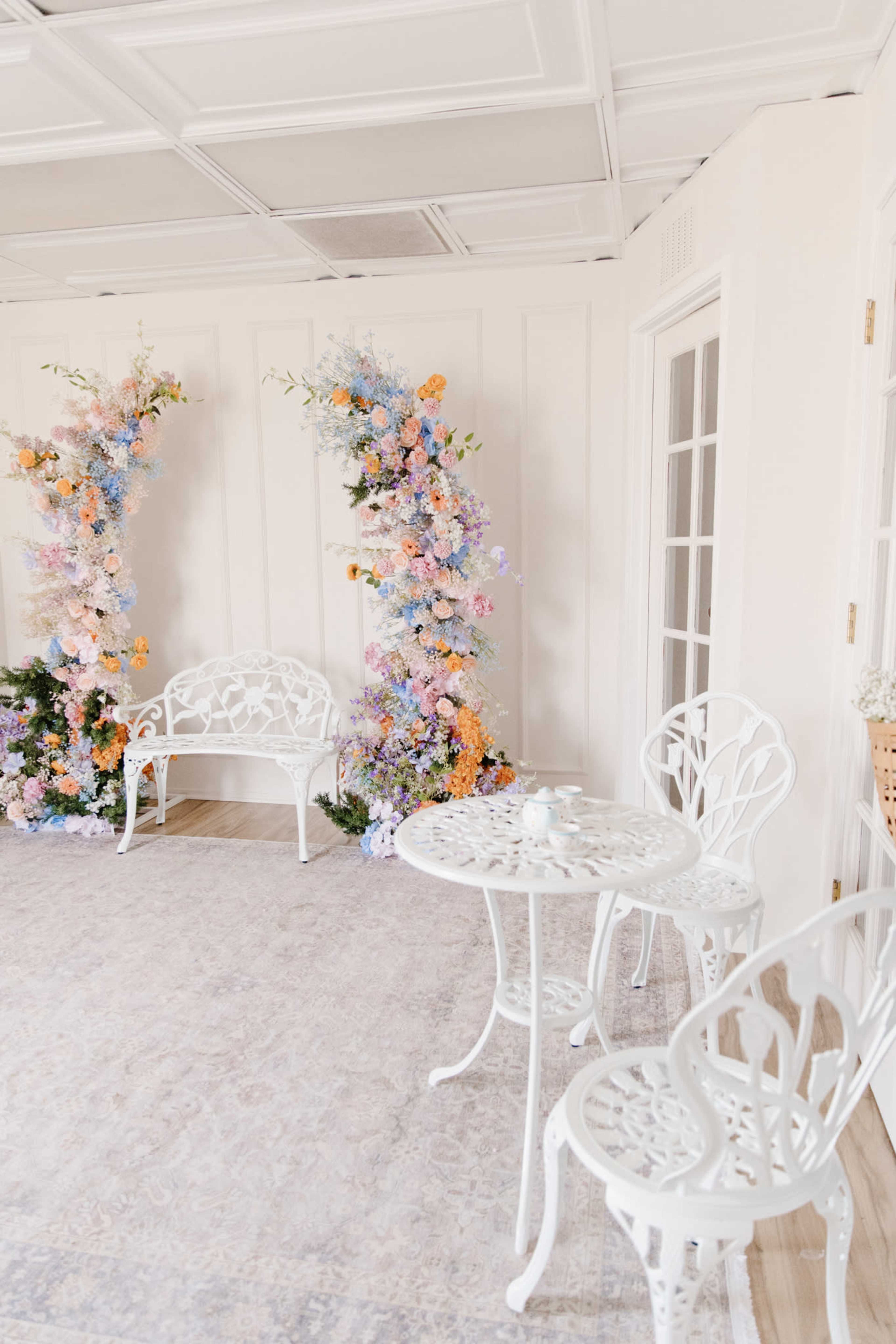 The image shows a light and airy room with two floral arrangements and a white iron table and chairs set on a patterned rug.