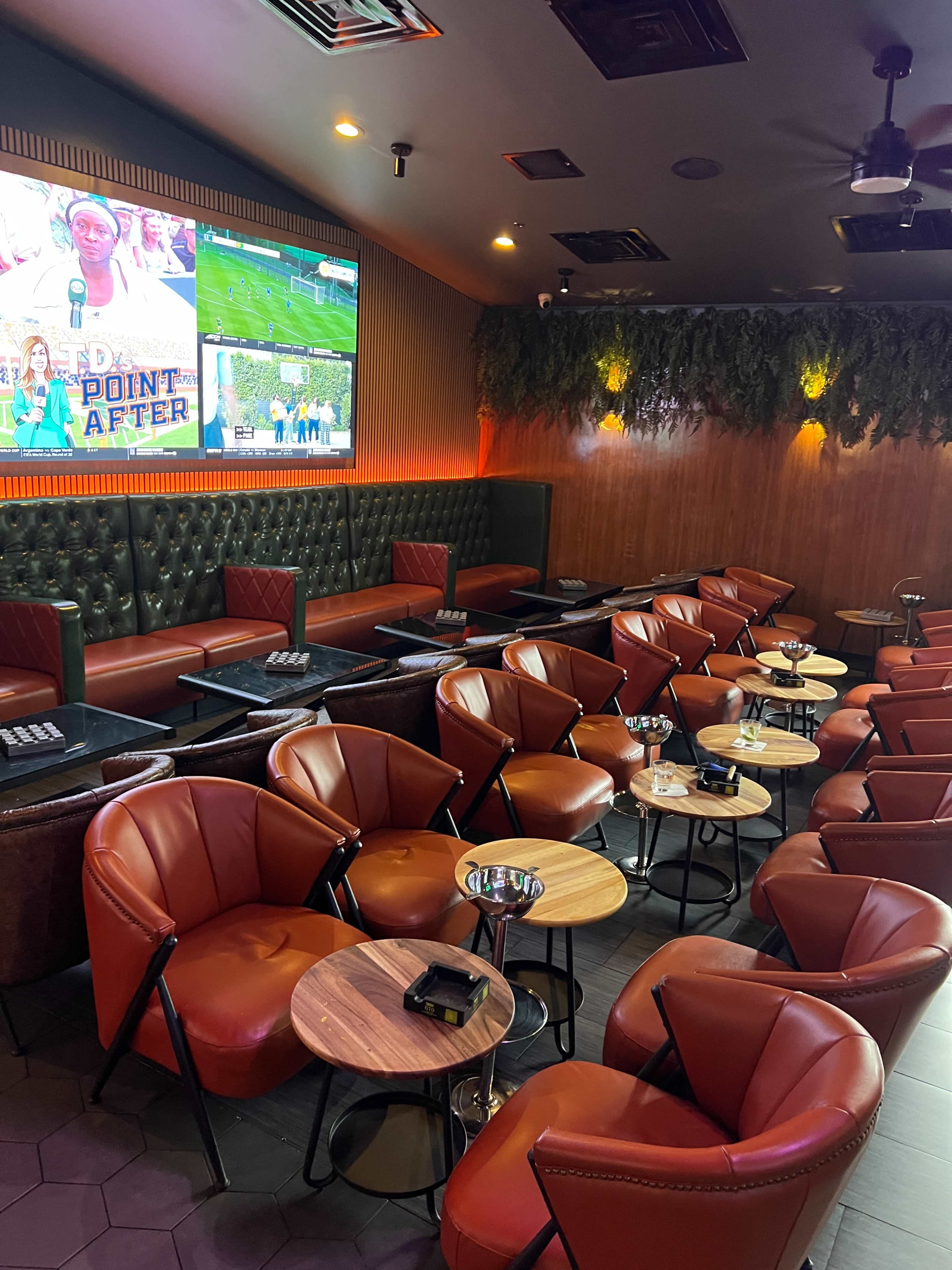 The image shows a modern lounge with red leather chairs and small circular tables, featuring a large screen displaying sports and green plants on the wall.