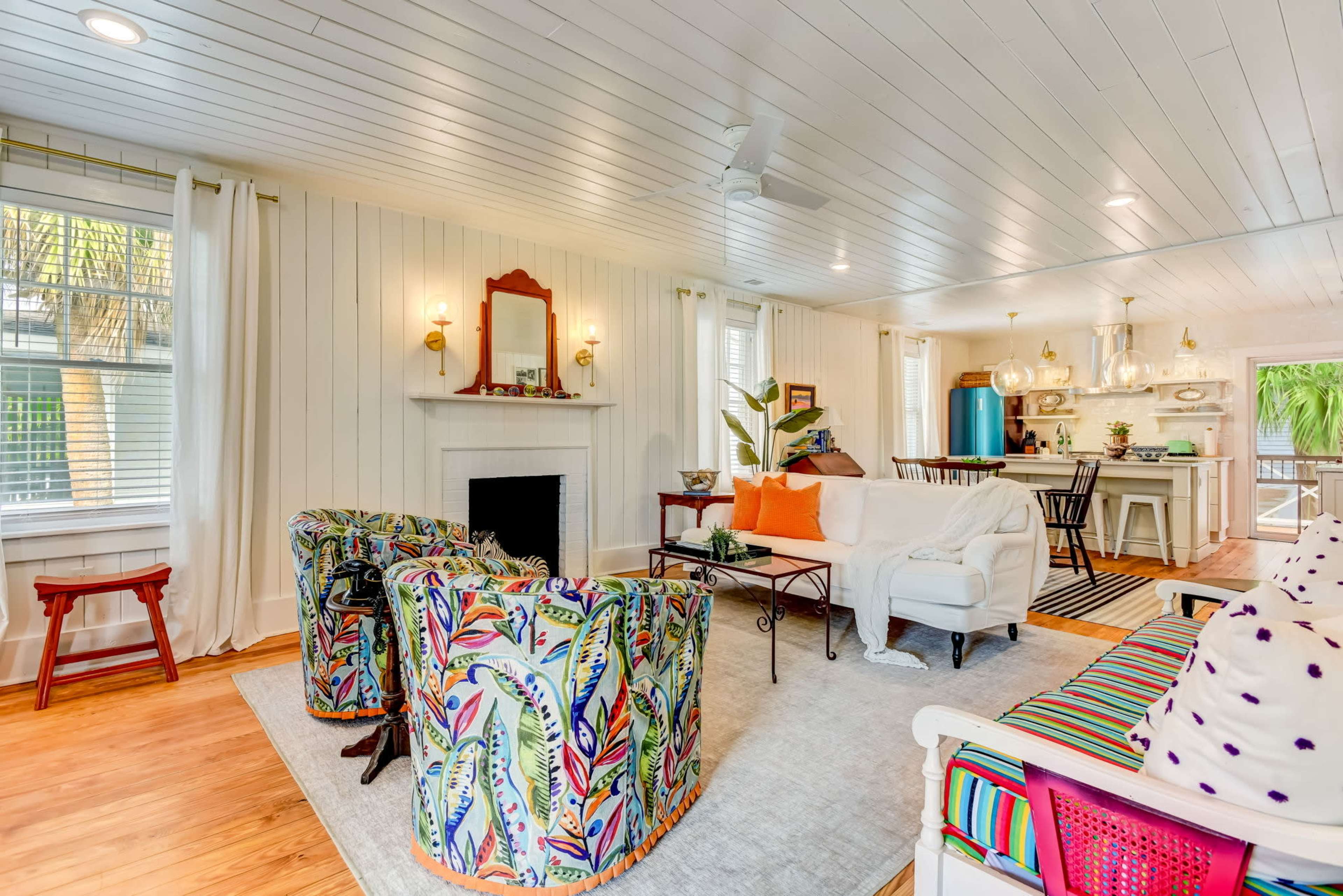 The image depicts a bright living room with colorful patterned chairs, a white sofa, a fireplace, and a dining area in the background.